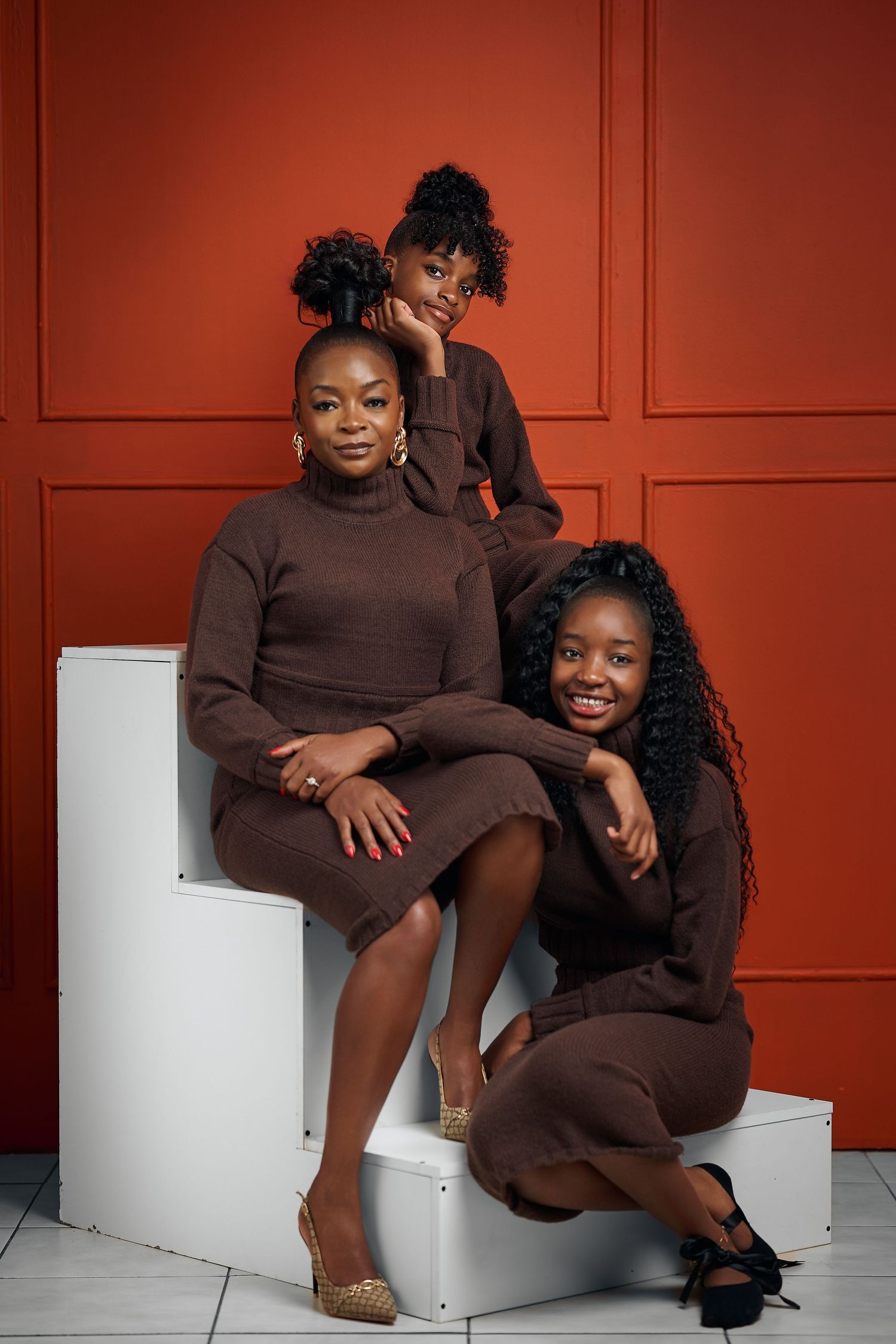 Three women in matching brown outfits pose together against a red wall in a stylish studio portrait.