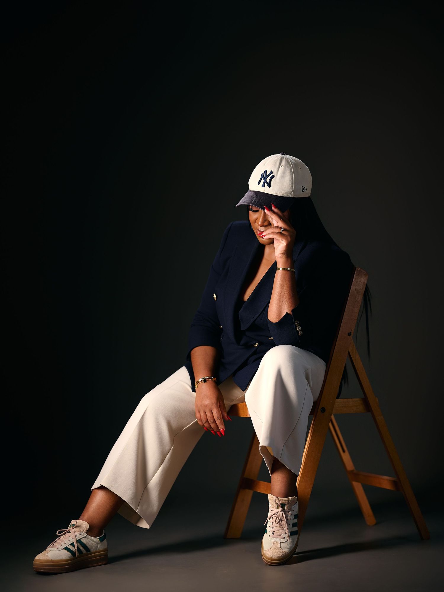 Person sitting on wooden chair wearing NY Yankees cap, navy blazer, white trousers, and sneakers in studio setting.