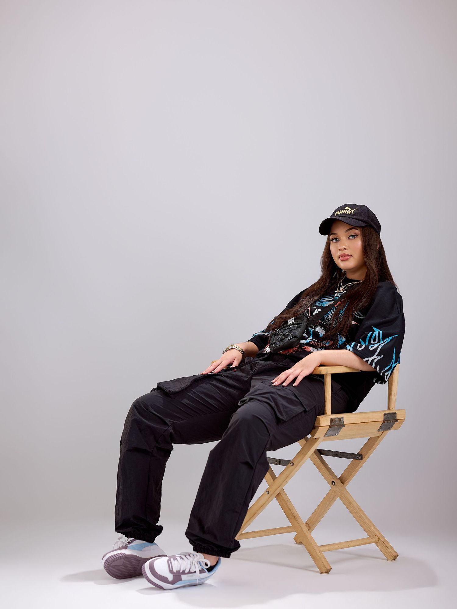 Young woman in black graphic shirt, cargo pants, and sneakers sitting on a wooden director's chair against gray background.