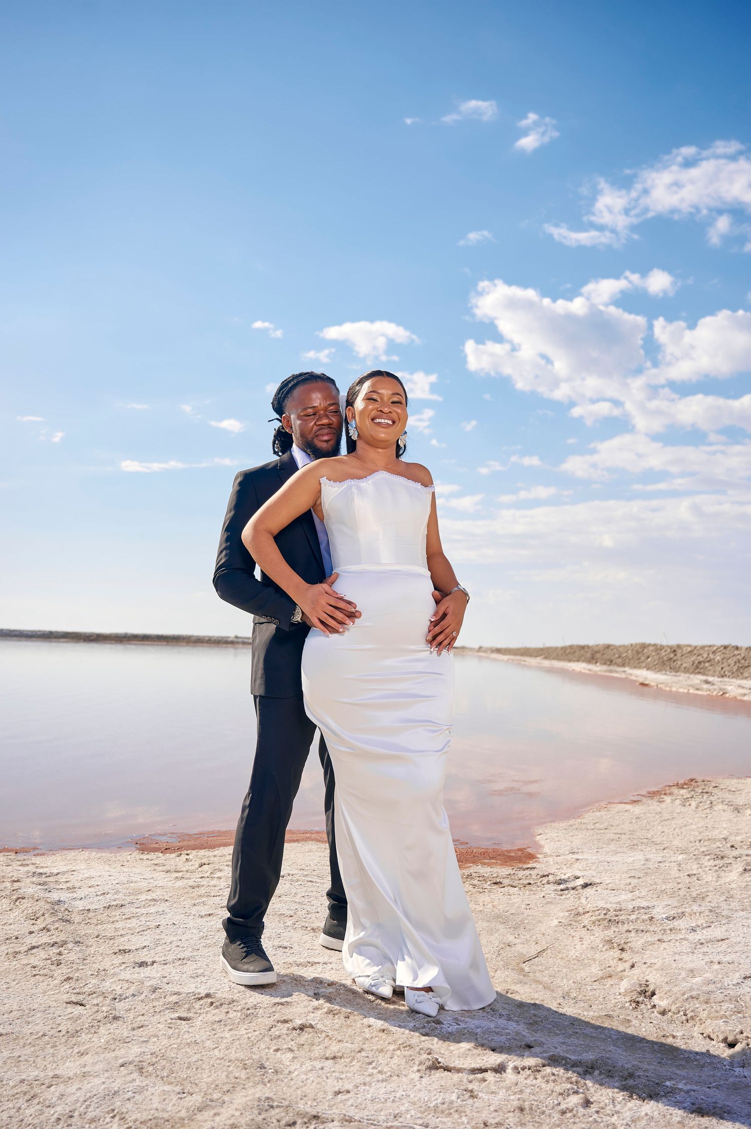 A joyful couple in wedding attire poses on a sandy shore beside a pink lake under a bright blue sky.
