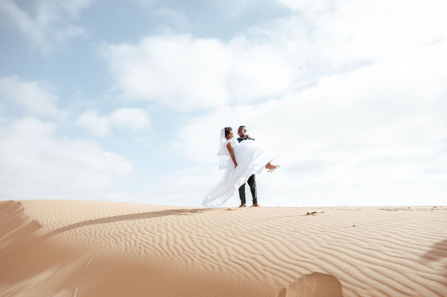 Couple in wedding attire dancing on golden sand dunes under a blue sky in a desert landscape.