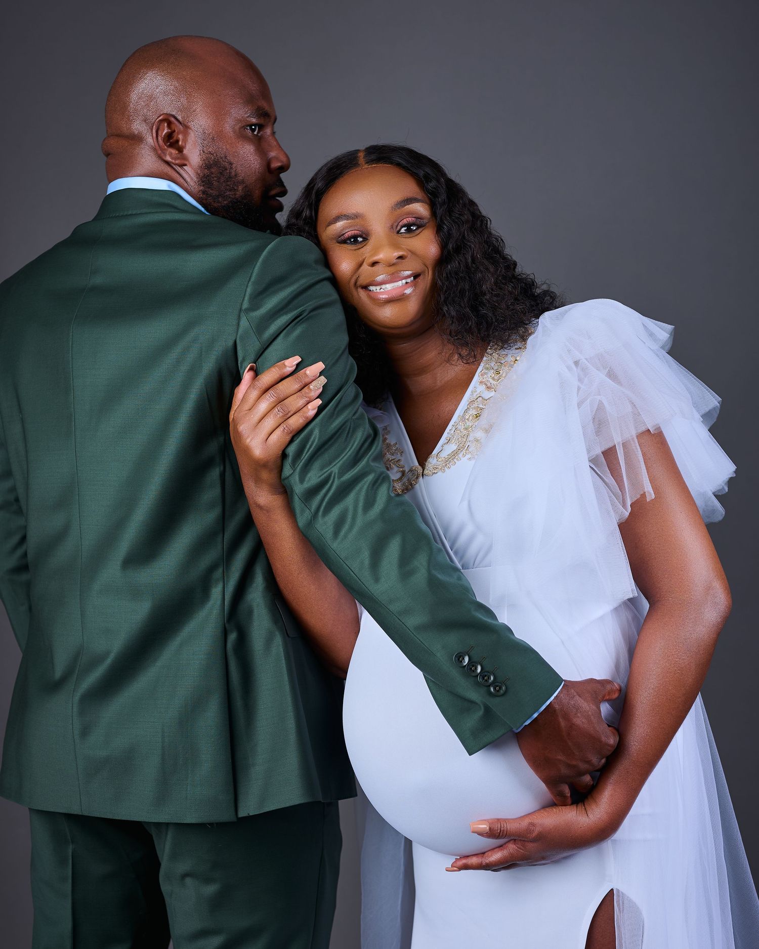 Pregnant woman in white dress poses with man in green suit during maternity photo shoot.