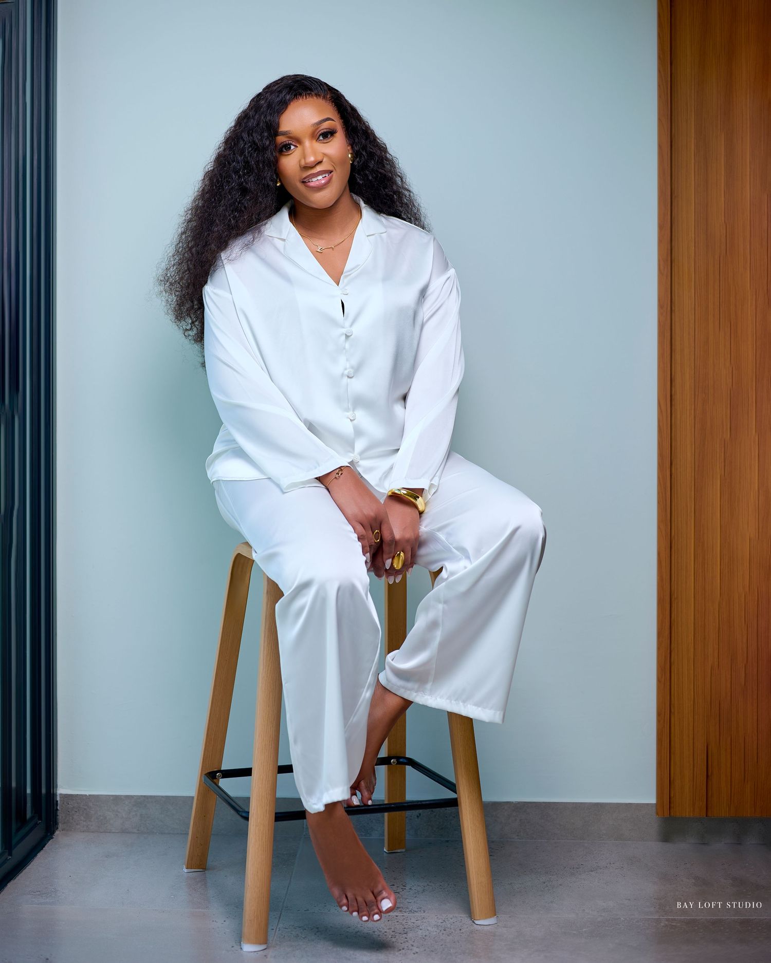 A person in crisp white loungewear sits barefoot on a wooden stool against a minimalist backdrop.