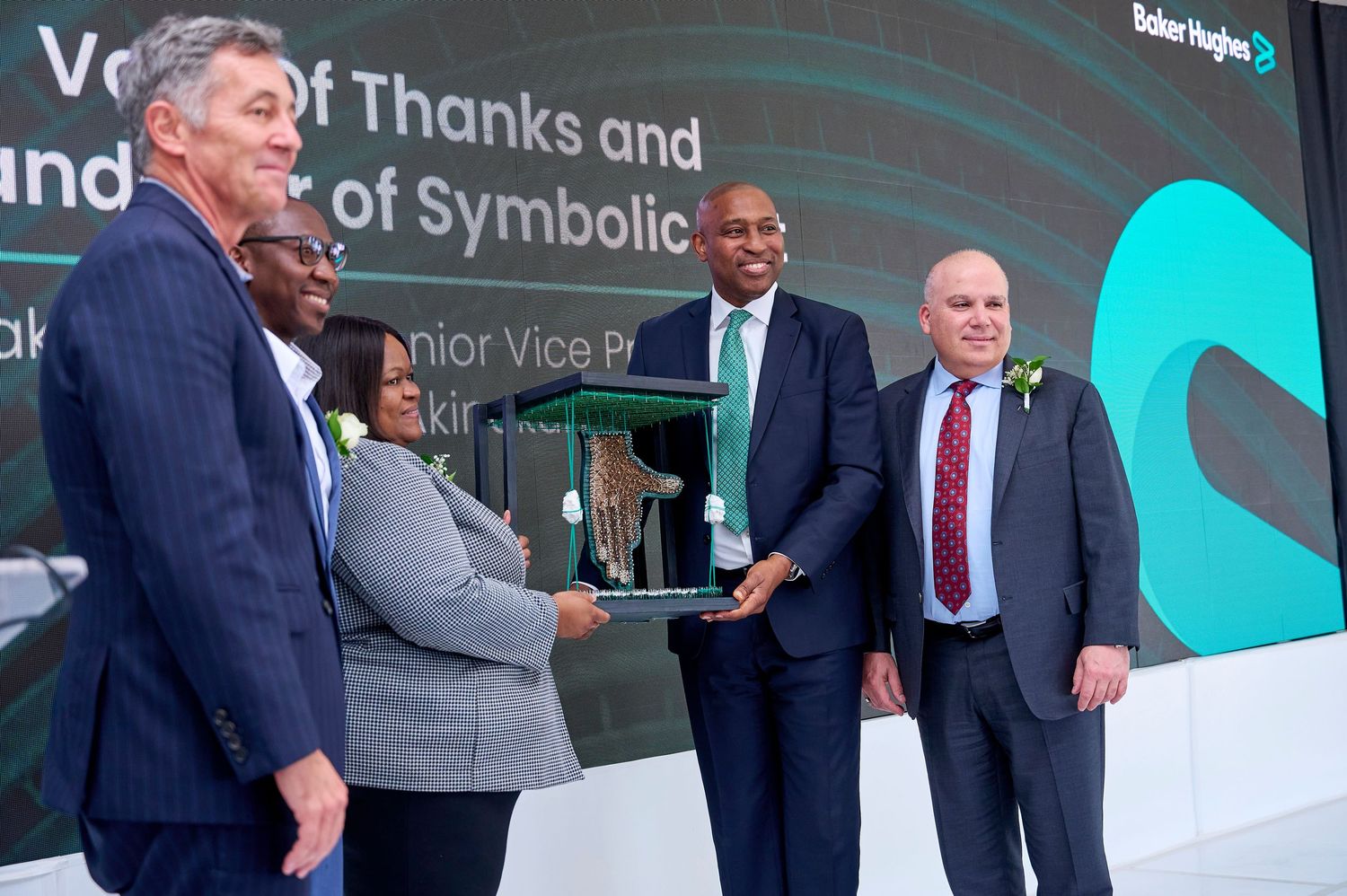 Baker Hughes executives exchange symbolic award at a formal ceremony, with attendees in business attire on stage.