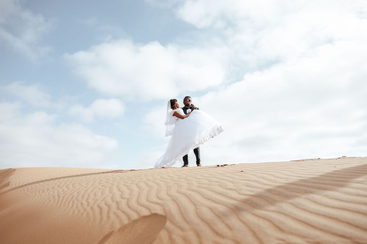 Couple in white wedding attire dancing on golden sand dunes under a cloudy sky in a desert setting.