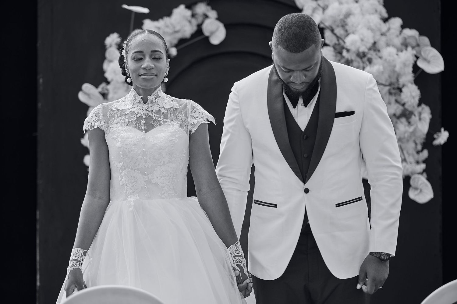 Black and white wedding photo of bride in lace gown and groom in white tuxedo holding hands at altar.