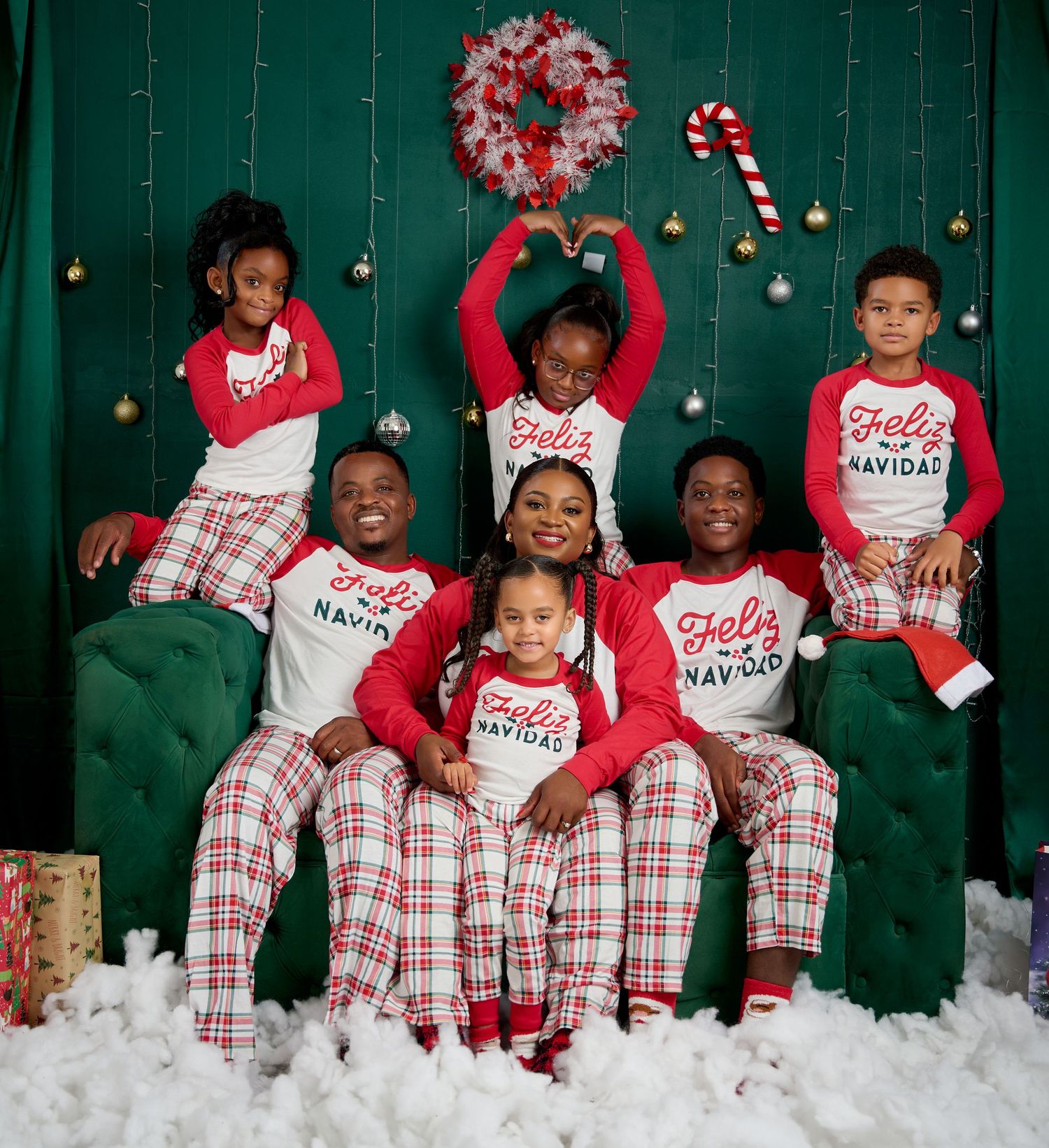 Family wearing matching red and white plaid Christmas pajamas pose together on green couch with holiday decorations.