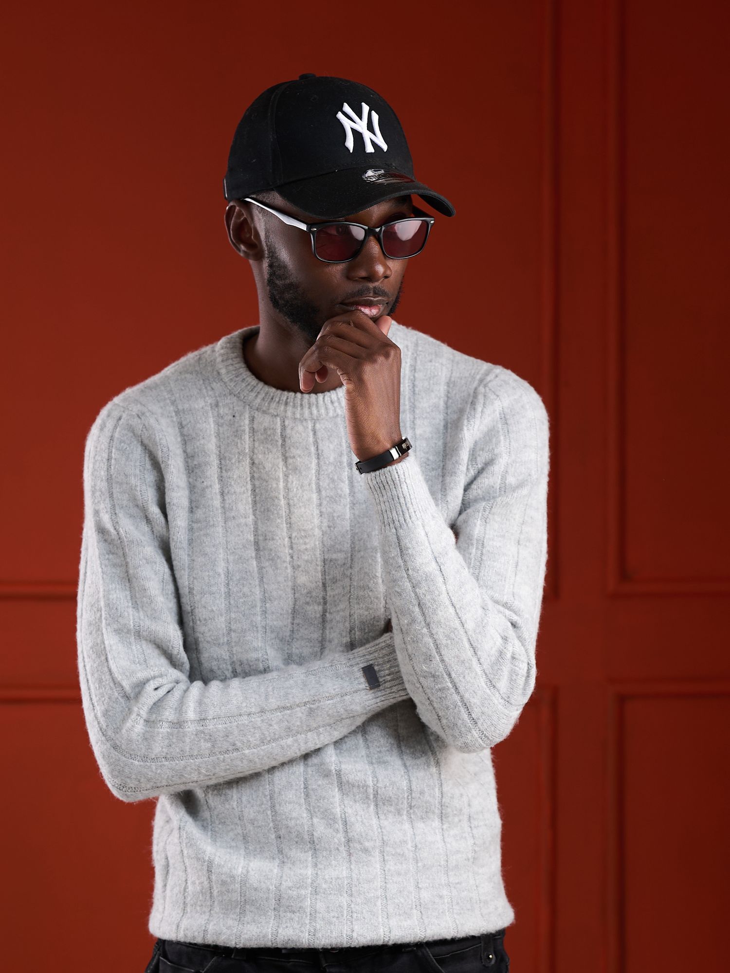 Man in gray ribbed sweater, NY Yankees cap, and red sunglasses posing thoughtfully against a dark red background.