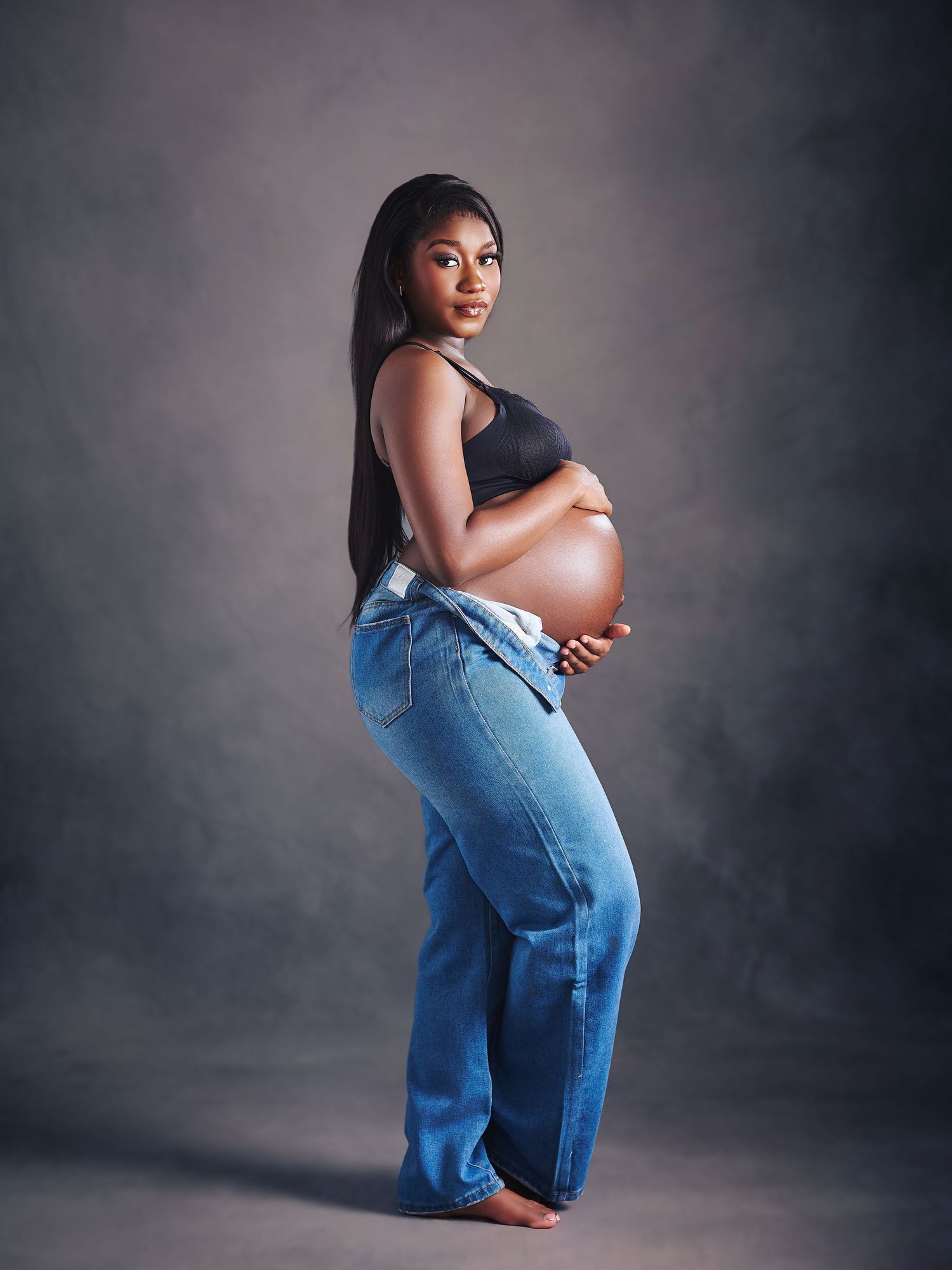 Pregnant woman in black crop top and open blue jeans holding baby bump, posing for maternity photo shoot.