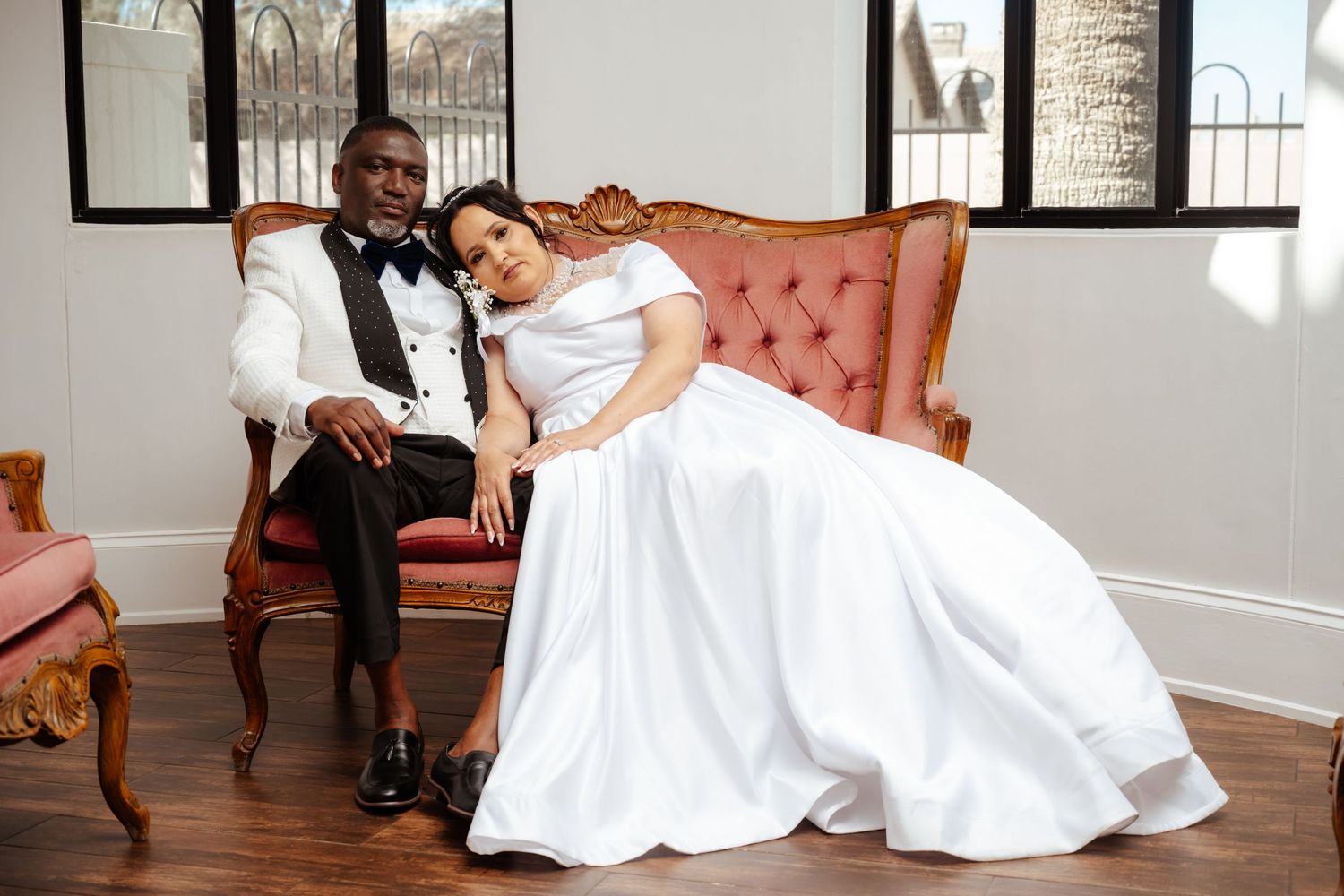 Elegant interracial wedding couple posing on a vintage pink velvet sofa, groom in white tuxedo, bride in flowing white gown.