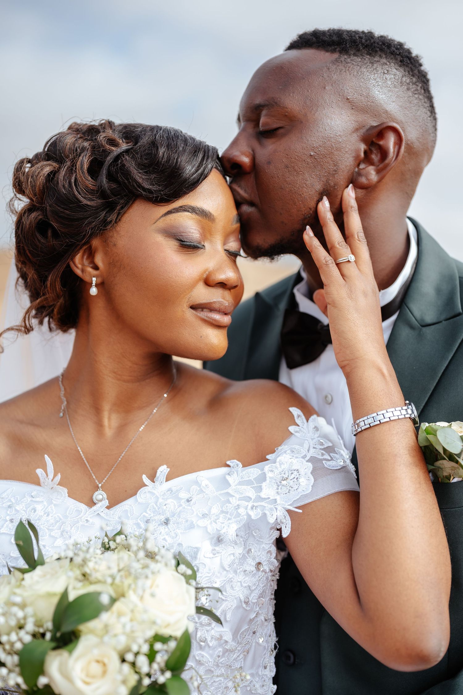 A groom in a green suit kisses his bride on the cheek as she smiles holding white roses in a lace wedding gown.