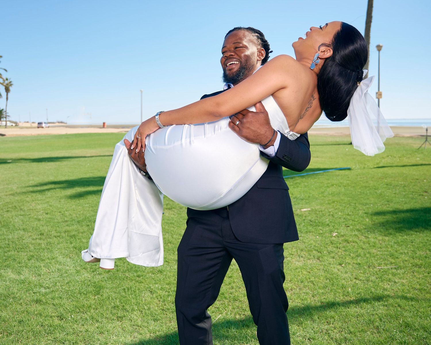 Groom in black suit lifting bride in white dress outdoors on green lawn with blue sky background.