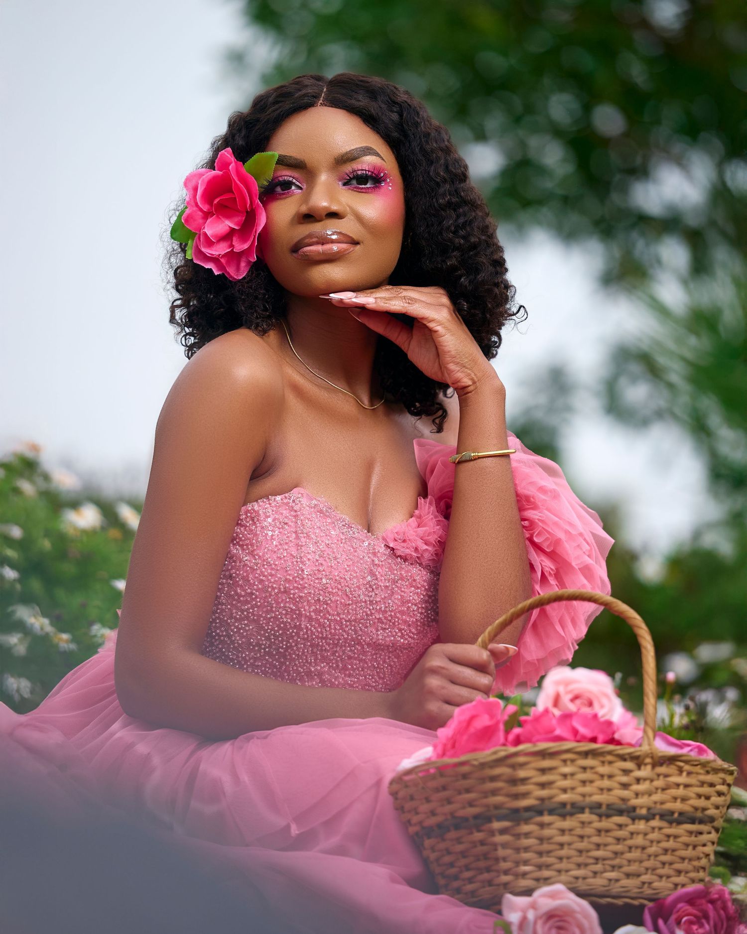 A woman in a pink tulle gown poses gracefully outdoors with a woven basket and floral accessories.