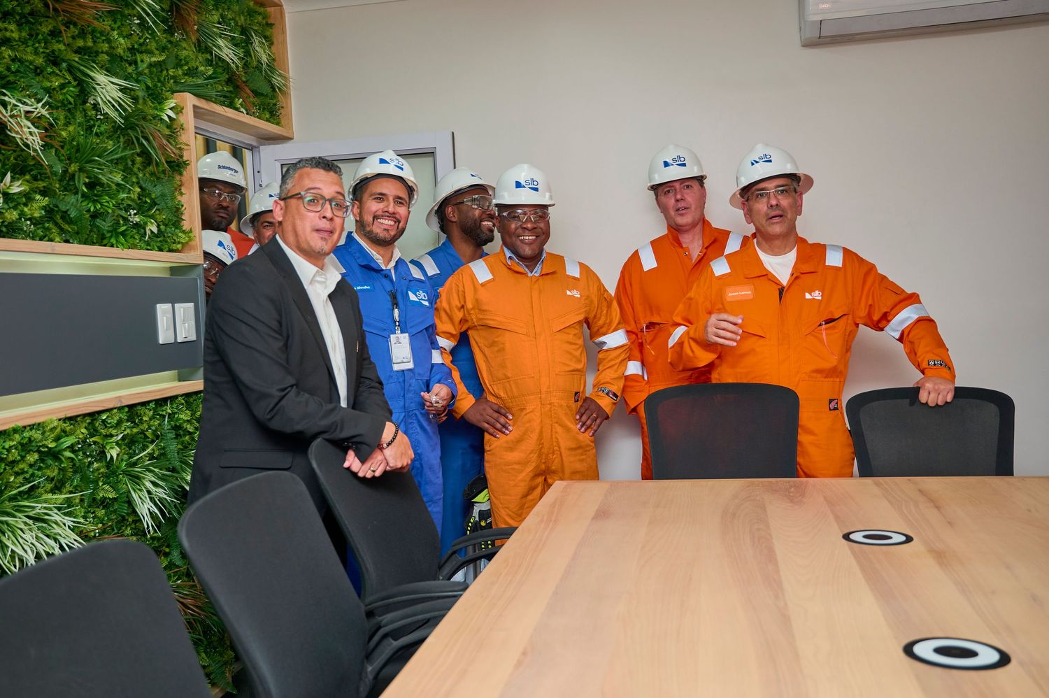 Group of workers in orange coveralls and helmets pose with two executives in a modern conference room.