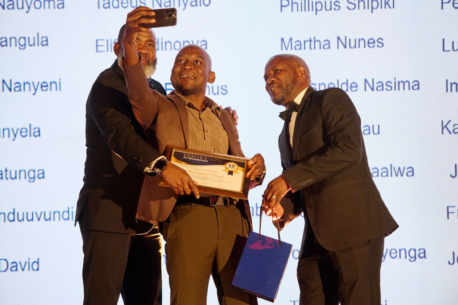 Three men celebrating award ceremony achievement holding certificate and gift bag on stage.