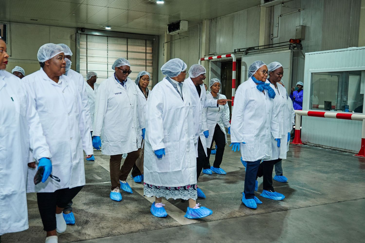 Group of professionals in white lab coats, hair nets, and blue shoe covers touring a food processing facility.