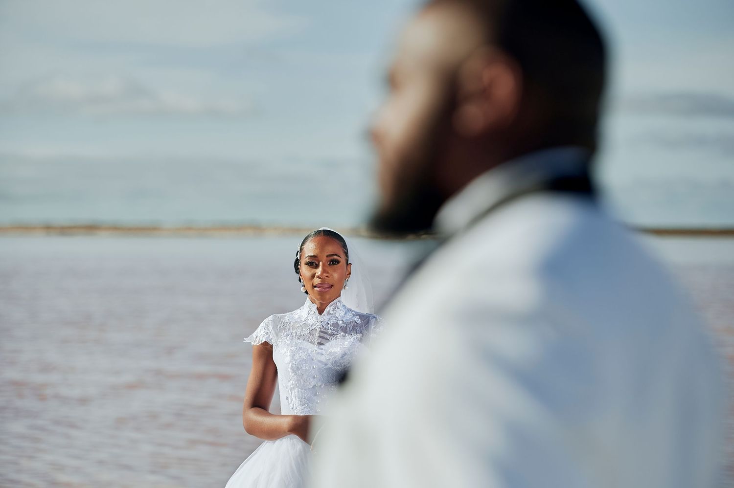 Bride in white lace gown stands by water as groom in white suit poses in foreground at outdoor wedding.