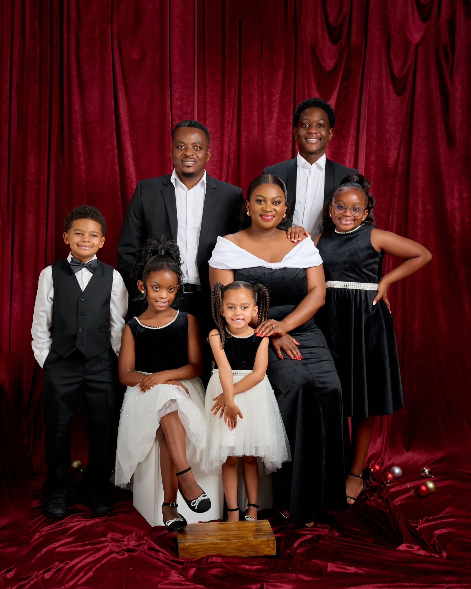 Family of seven posing elegantly in black and white formal attire against a rich red velvet backdrop.