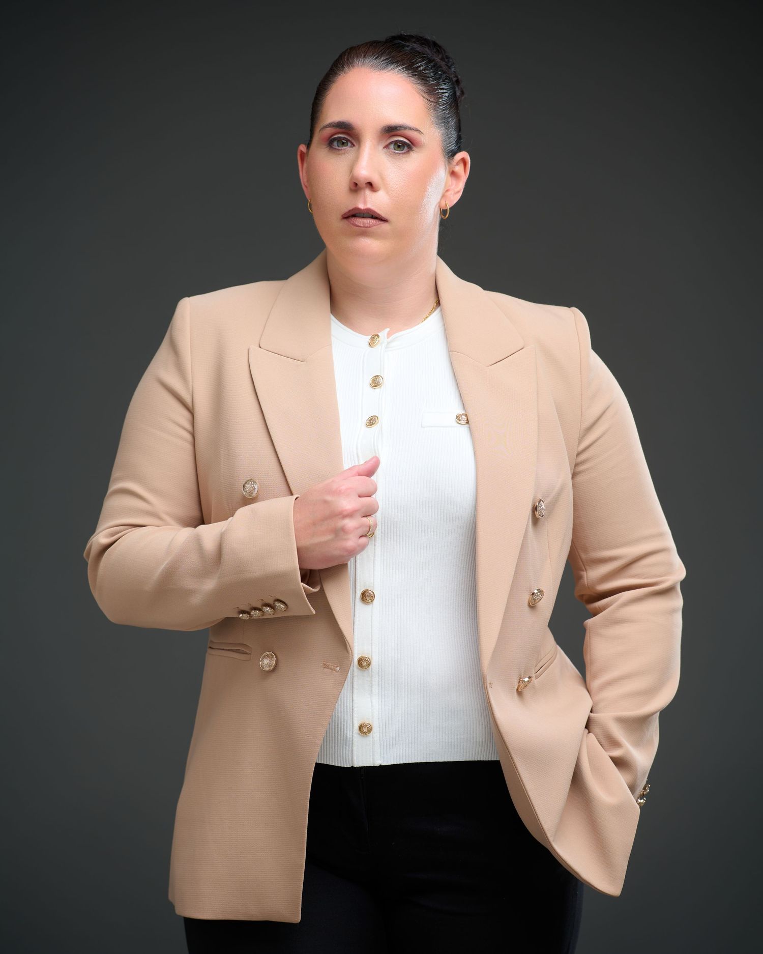 Professional studio portrait of someone wearing a tan blazer and white top against a dark background.