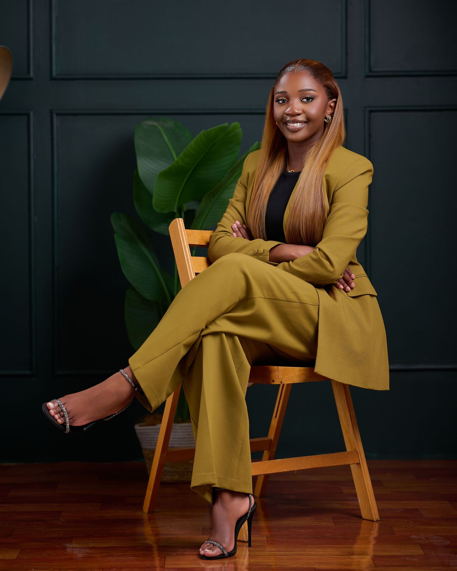 A woman in a stylish olive pantsuit poses confidently in various positions on a wooden chair against a dark wall.