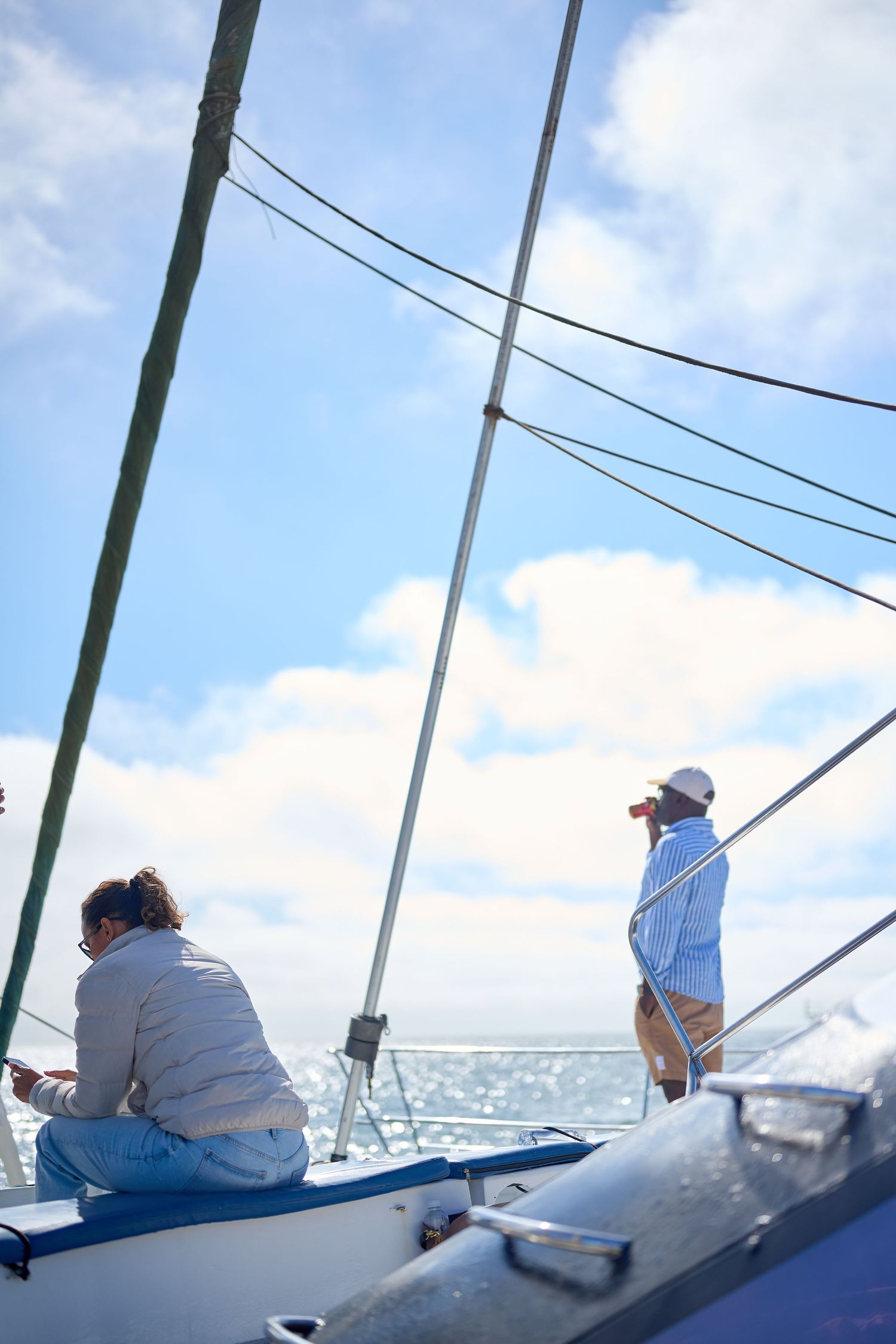 Two people relaxing on a sailboat deck under a bright blue sky with clouds, one sitting and one standing near the mast.