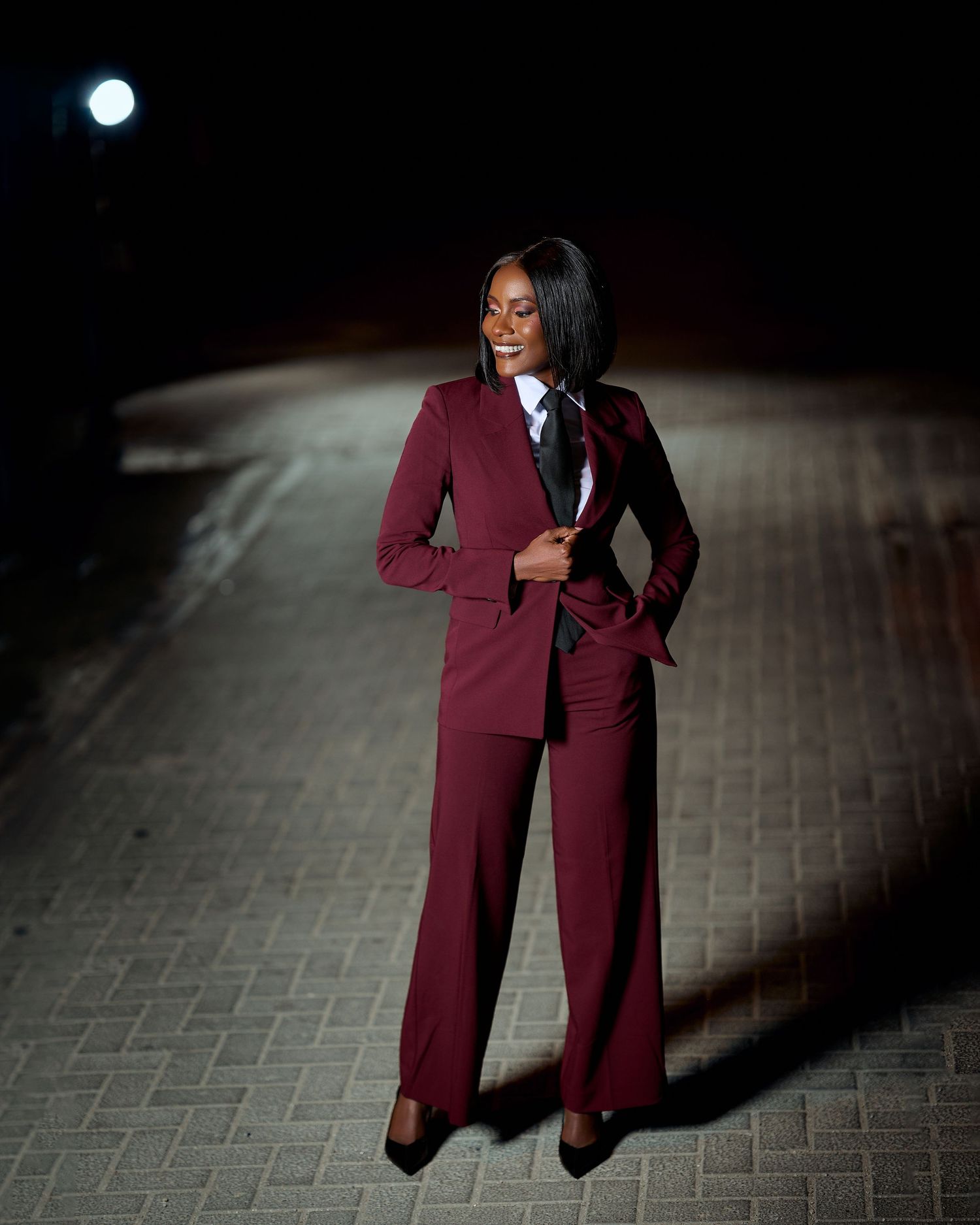 Woman in burgundy pantsuit posing confidently on a cobblestone path at night under bright light.