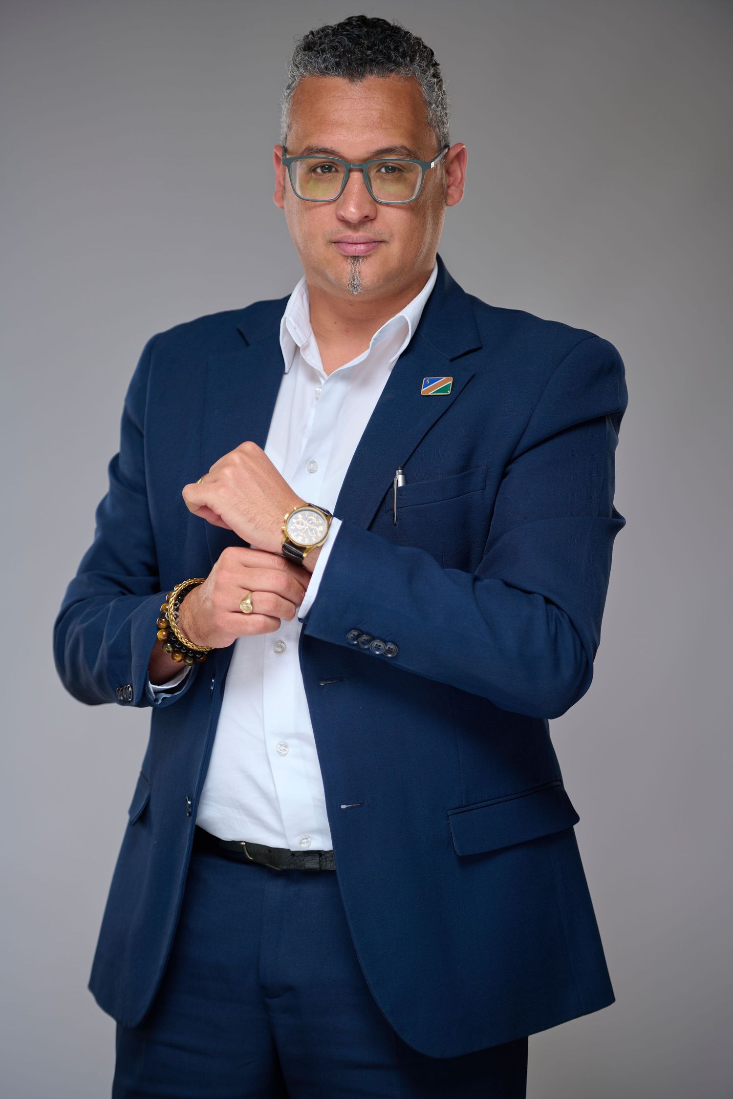 Professional man in navy blue suit adjusting cufflinks, wearing glasses and a gold watch, posing confidently against gray background.