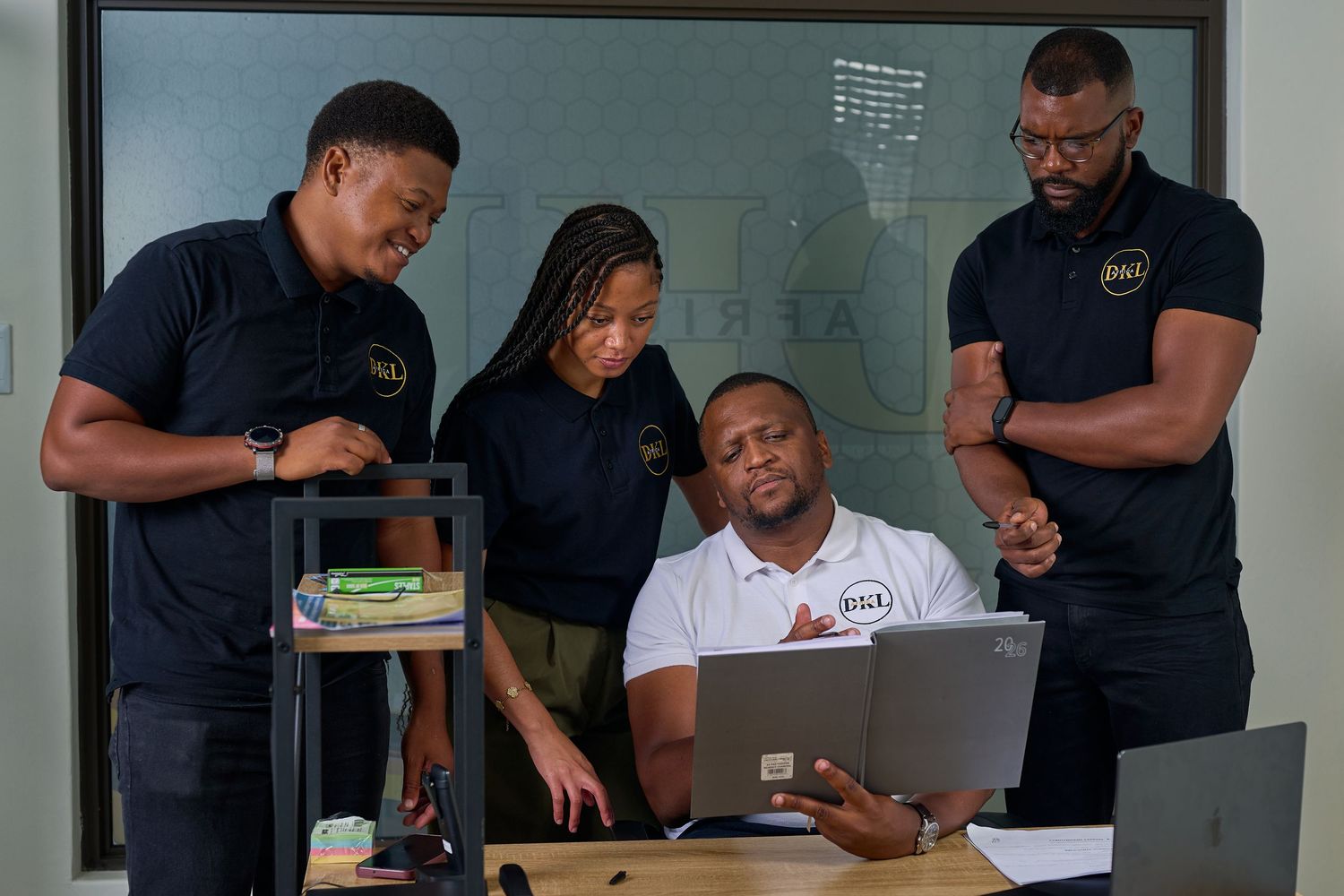 Four professionals in matching black uniforms collaborate around a laptop in a modern office setting.