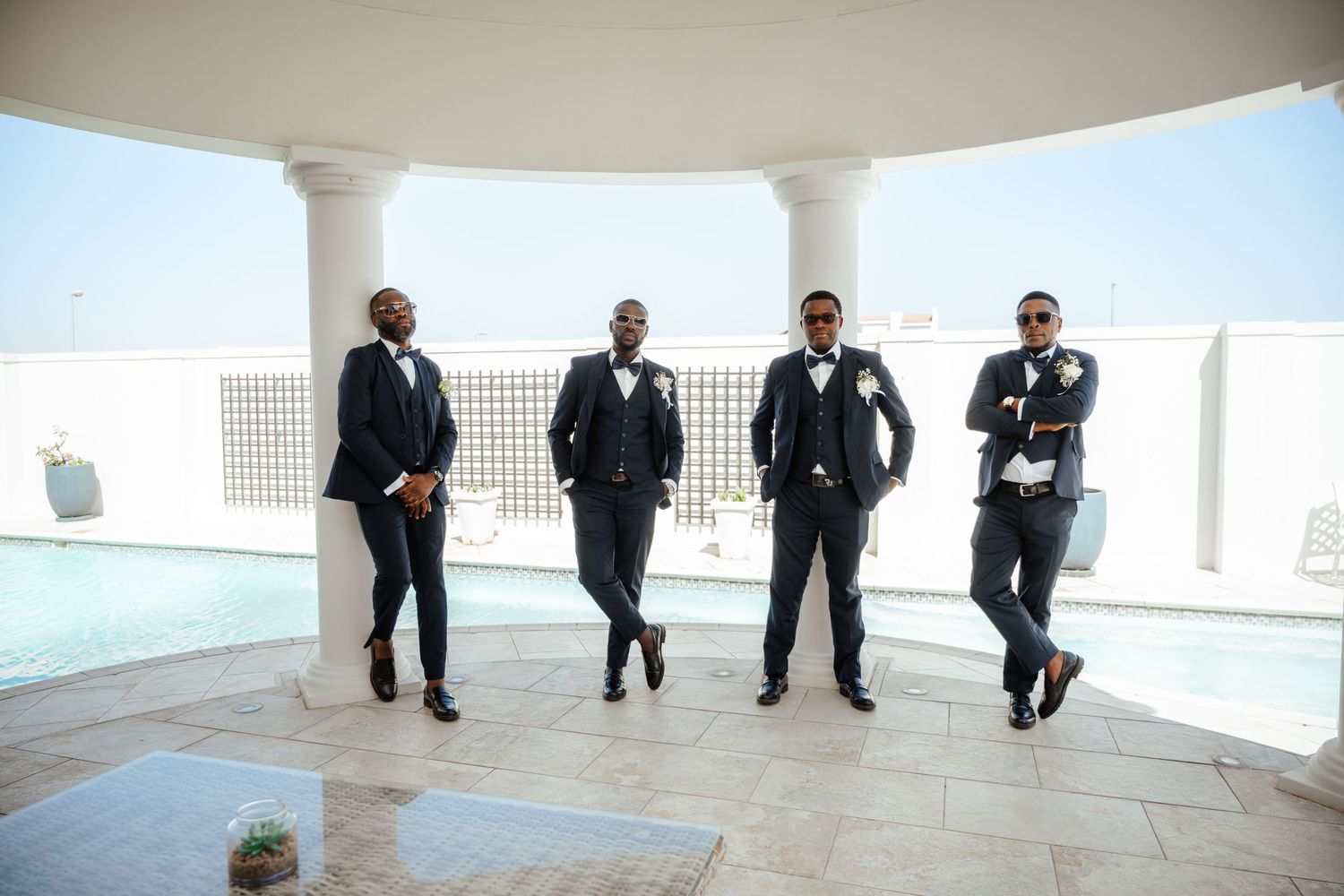 Four groomsmen in navy suits pose confidently on a rooftop terrace with white columns and a pool backdrop.