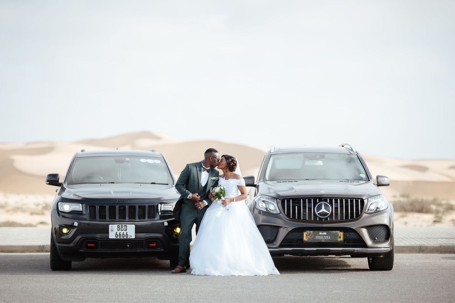 A bride and groom share a kiss between a black Jeep and silver Mercedes SUV in a desert landscape.
