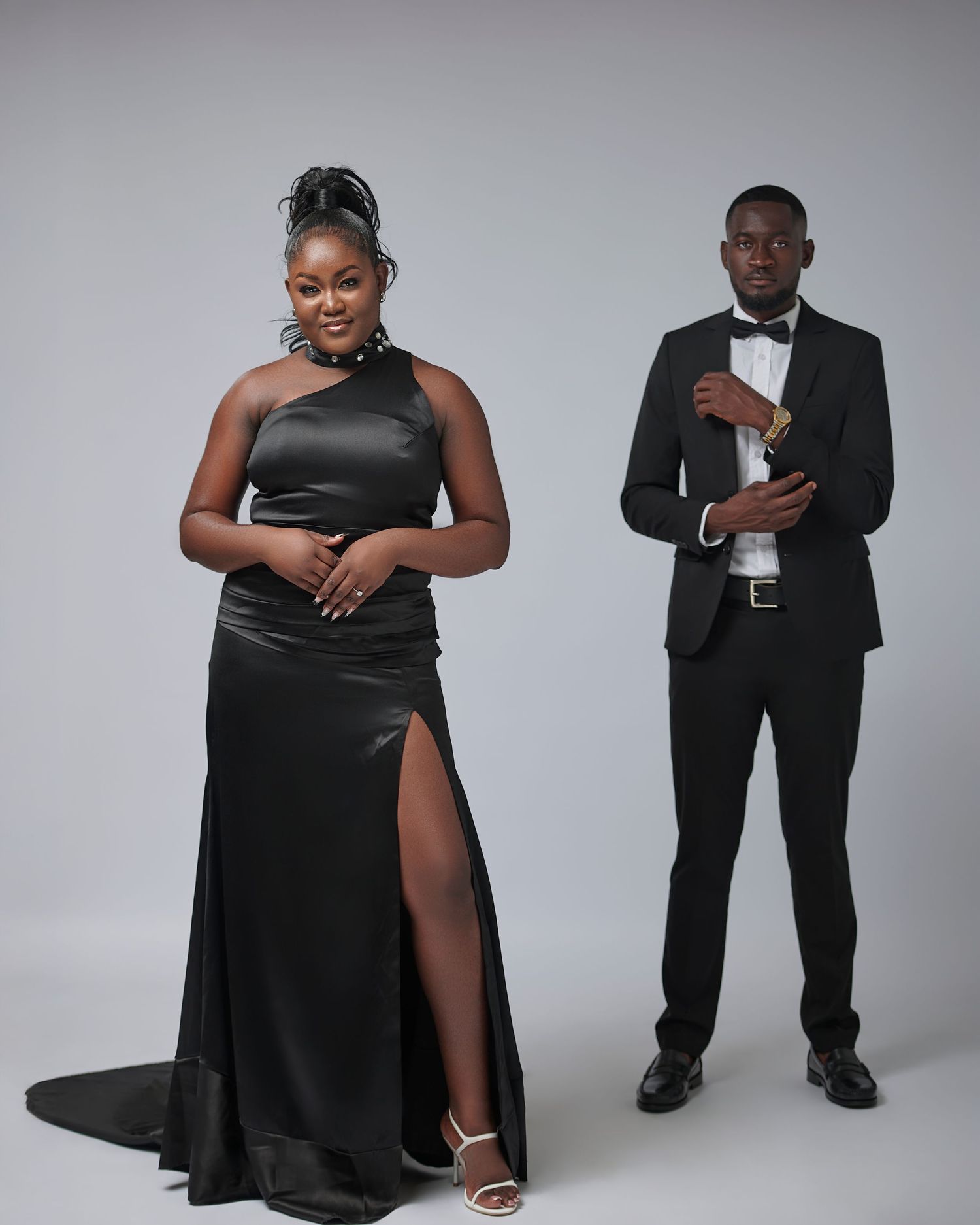 Elegant couple posing in matching black formal attire, woman in satin gown with slit, man in classic tuxedo with bow tie.