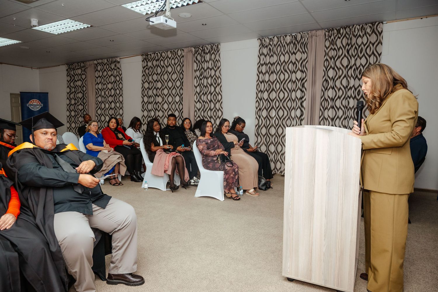 Woman speaking at podium during graduation ceremony with audience of graduates and guests seated in decorated hall.