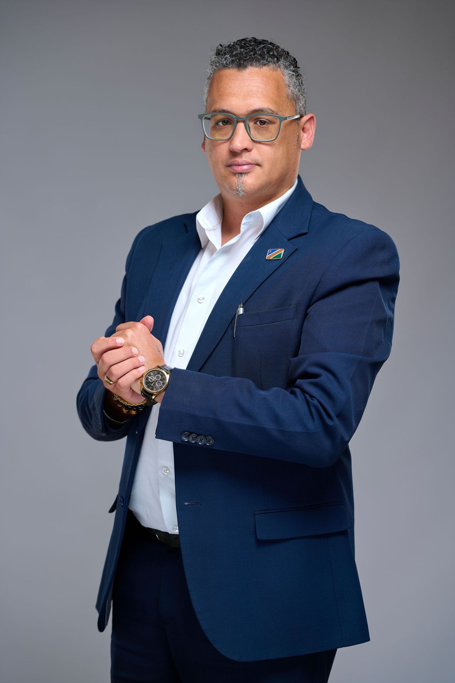 Professional man in navy blue suit with glasses posing confidently against gray background.