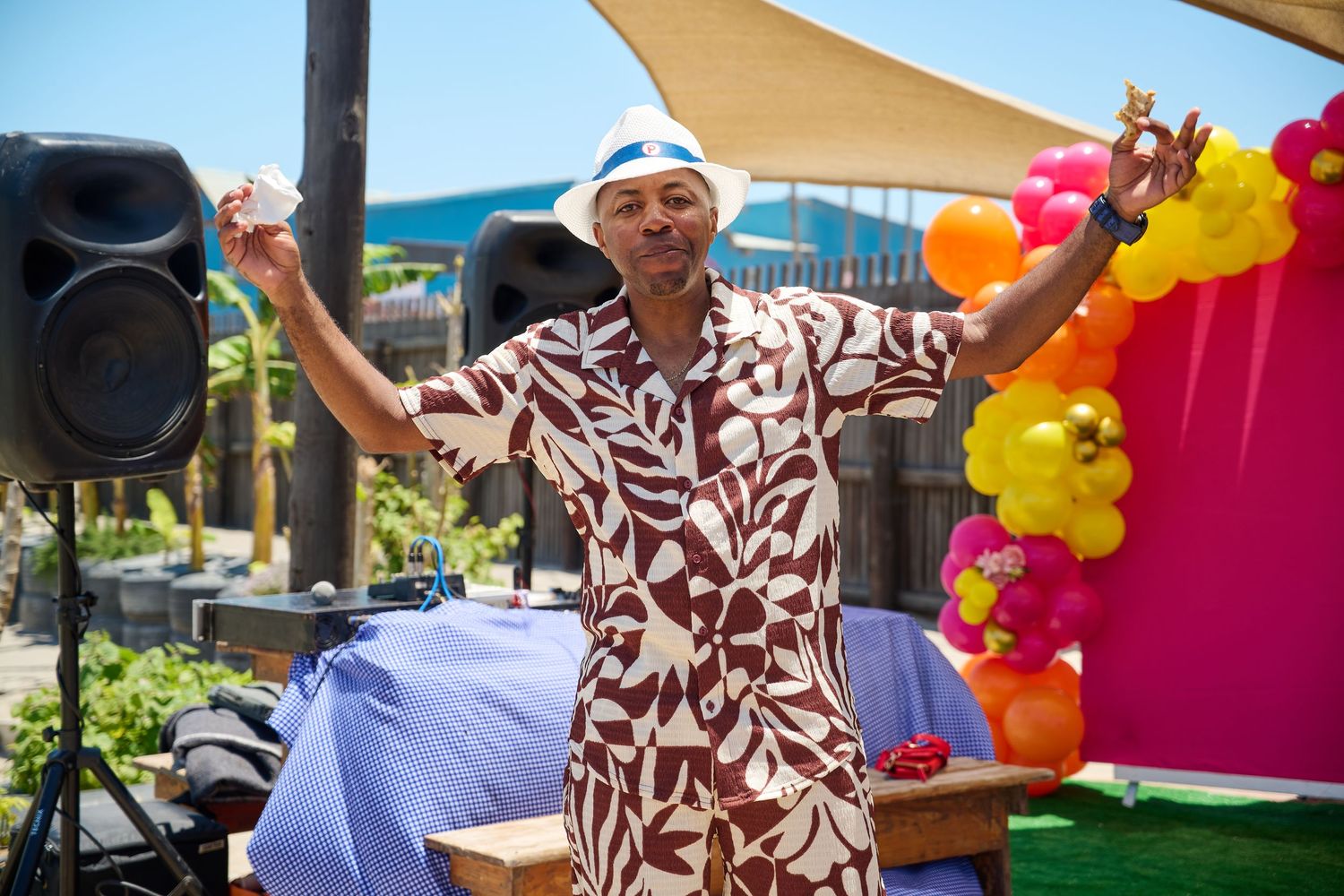 Man in floral shirt dancing at outdoor party with colorful balloon decorations and speaker on sunny day.
