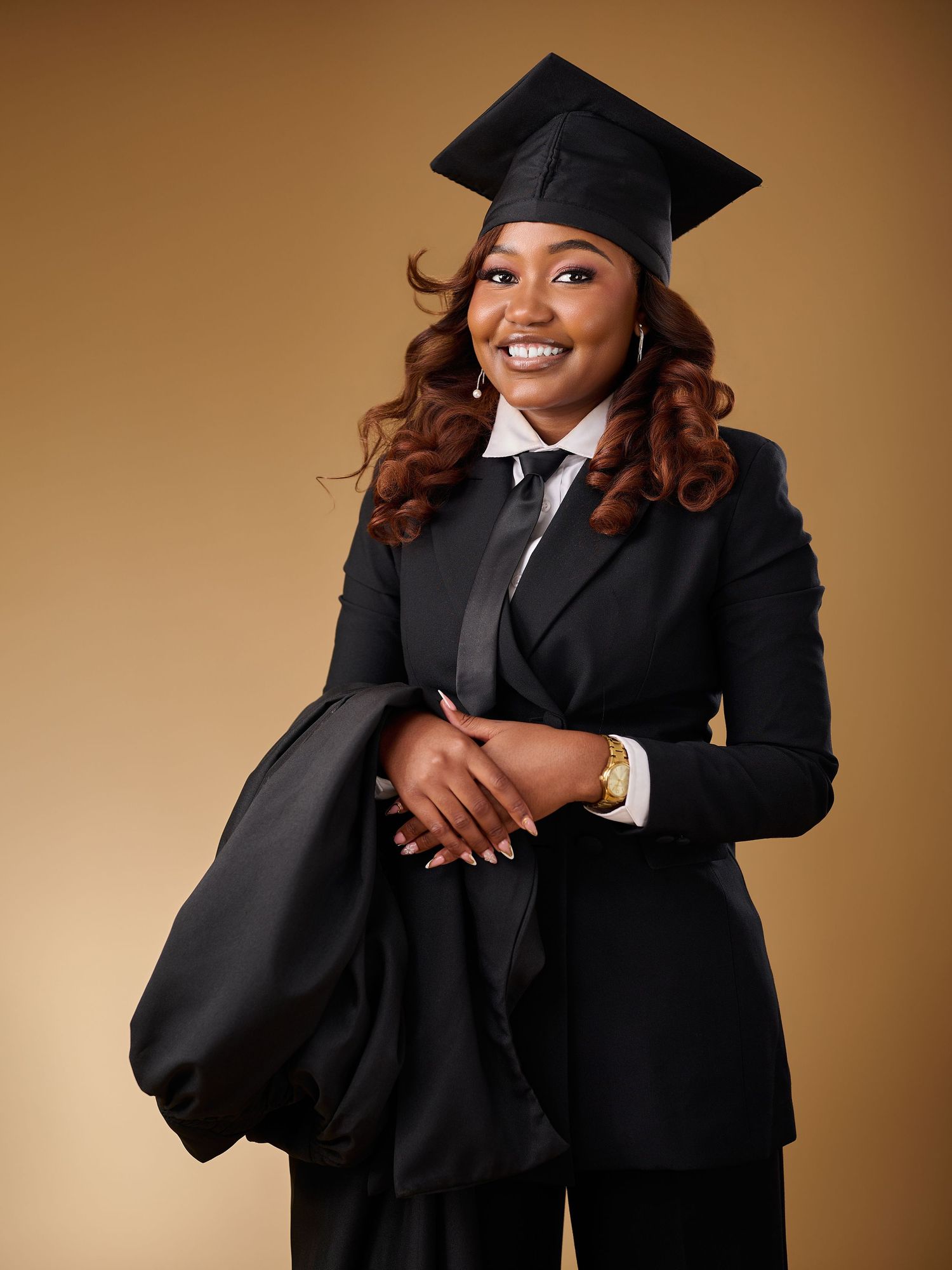Smiling graduate in black cap and suit holding academic gown against warm beige background.
