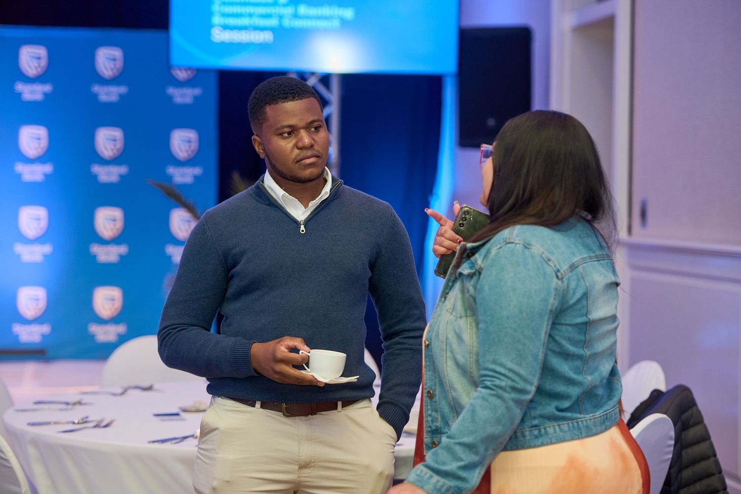 Two professionals networking at a corporate breakfast council session, man holding coffee cup in conference room.