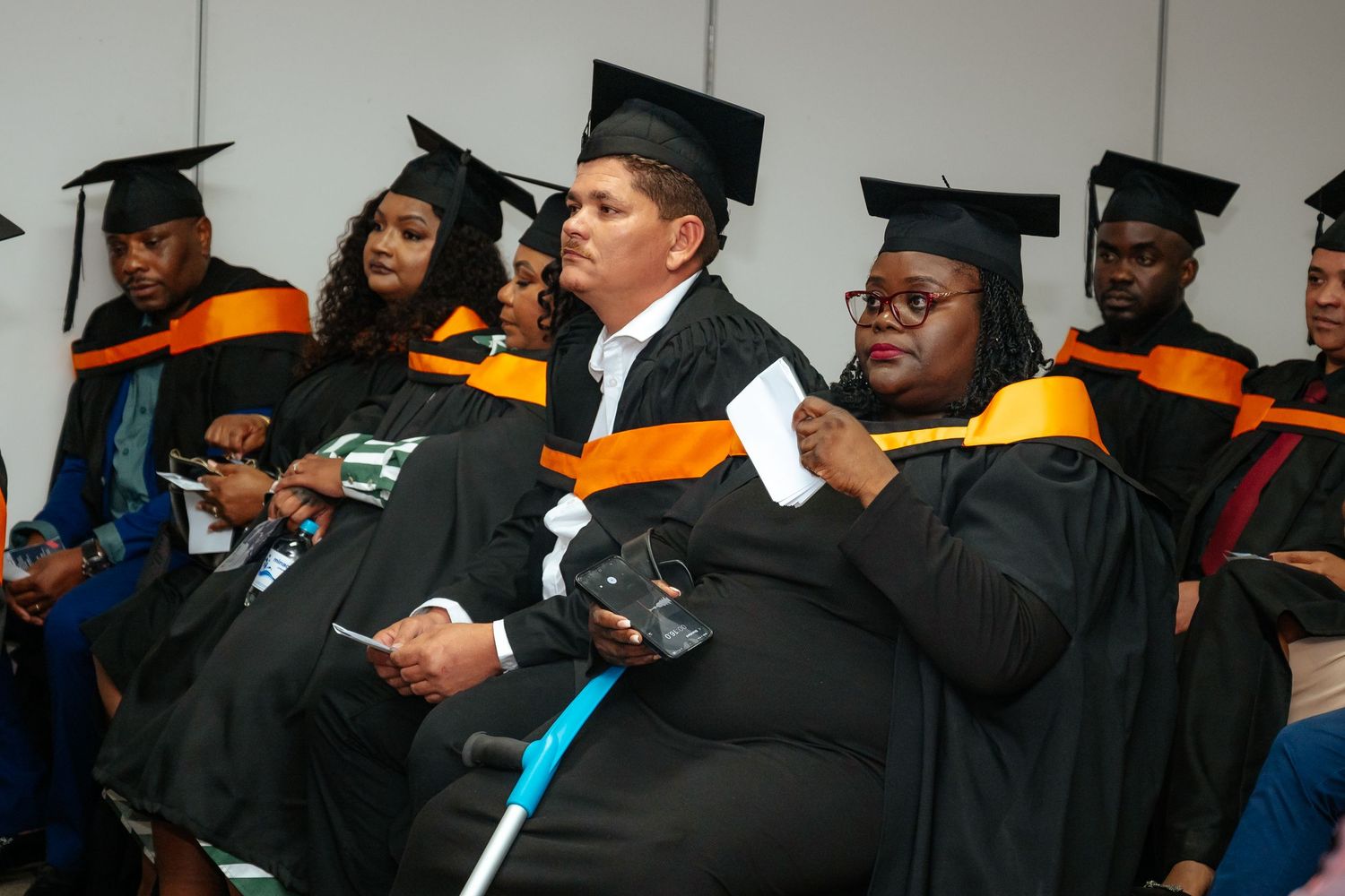 Graduates in black and orange academic regalia seated at a university graduation ceremony.