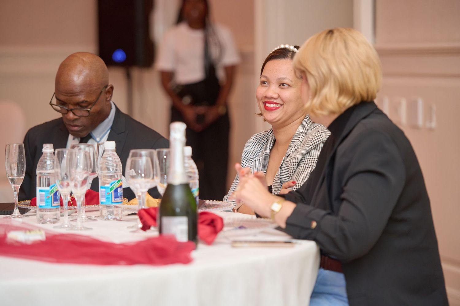 Three elegantly dressed guests smile and converse at a formal dinner event with wine glasses and bottled water on table.