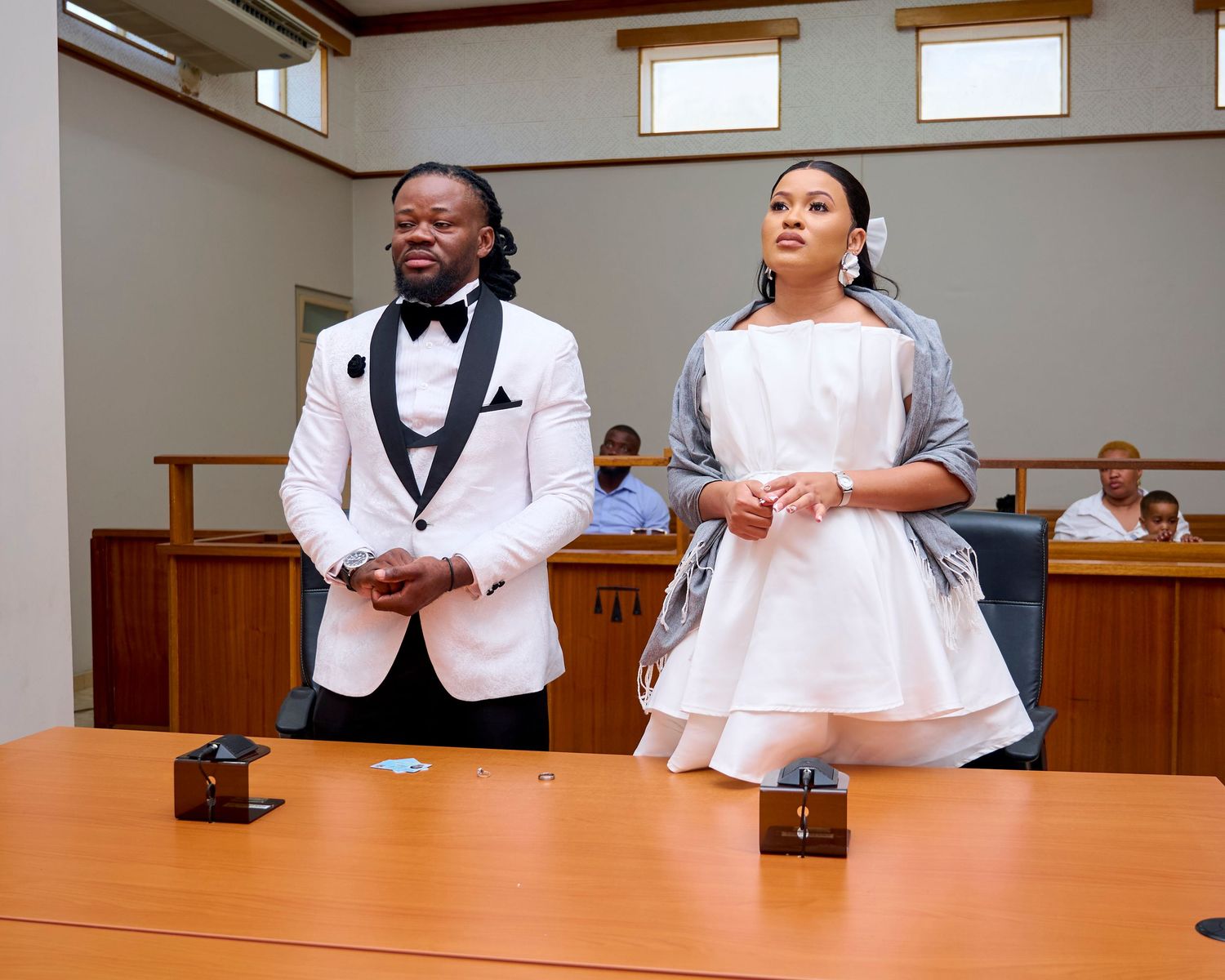 Couple in formal attire standing at a wooden table in a courtroom, man in white tuxedo, woman in white dress.