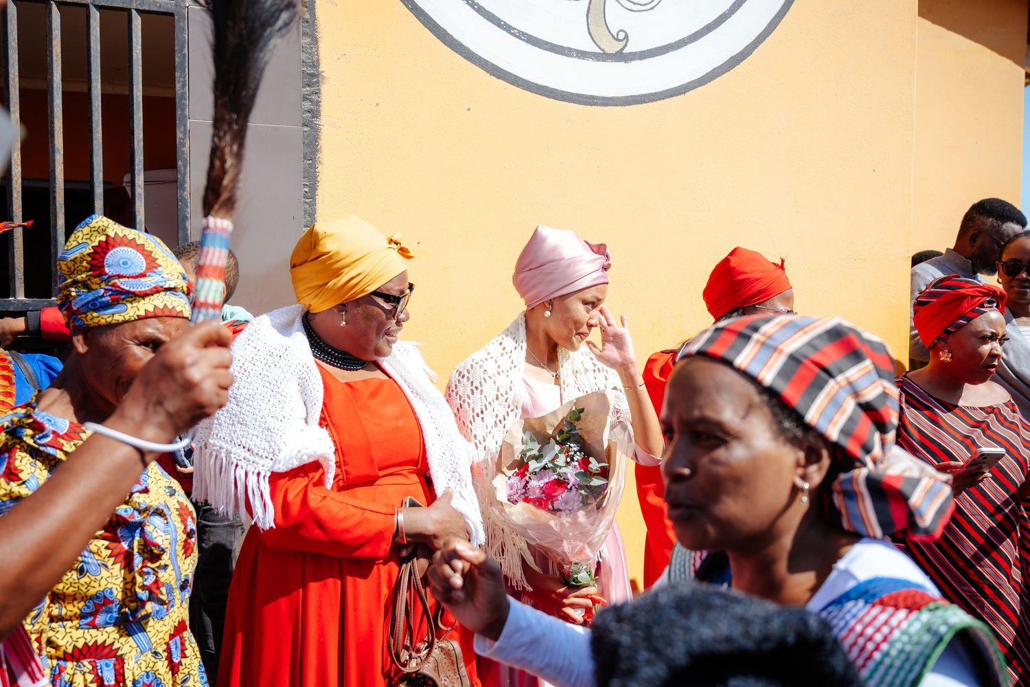Colorful African women in traditional attire and headscarves celebrate outdoors near an orange wall.