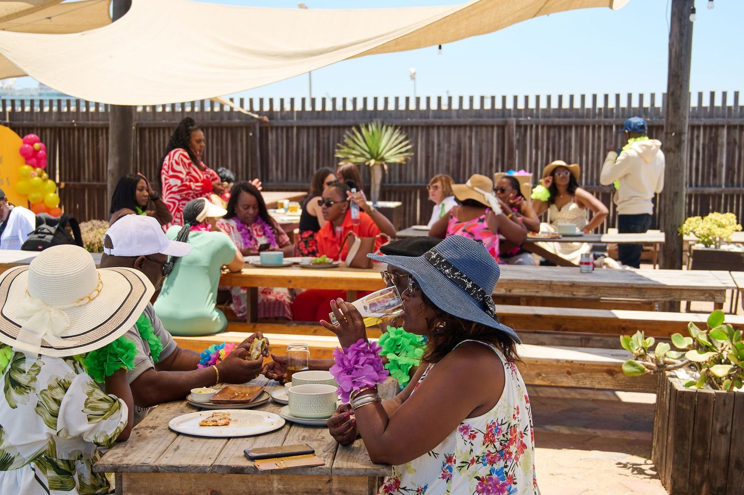Group of women in colorful outfits enjoying an outdoor party at a wooden picnic table.