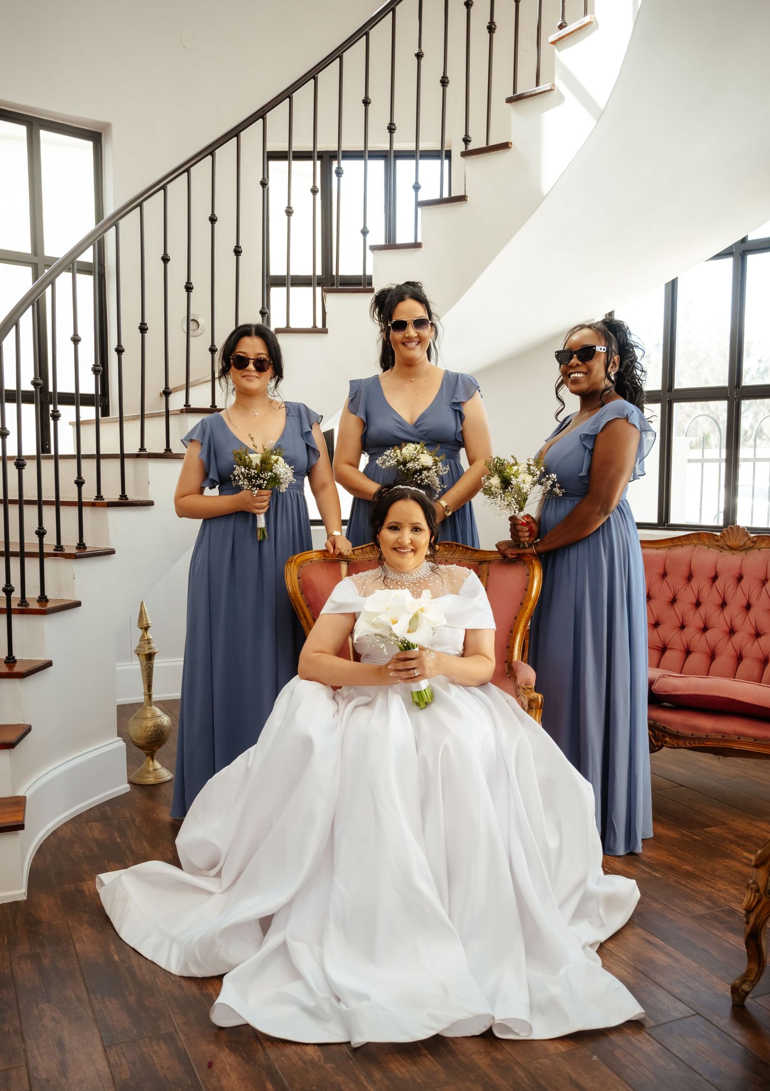 Bride in white gown seated with three bridesmaids in blue dresses holding bouquets near elegant staircase.