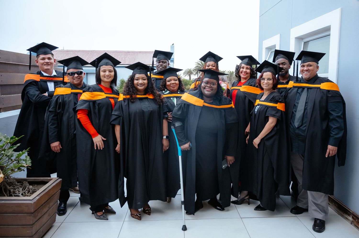 Group of graduates in black and gold academic gowns and caps posing together outdoors at graduation ceremony.