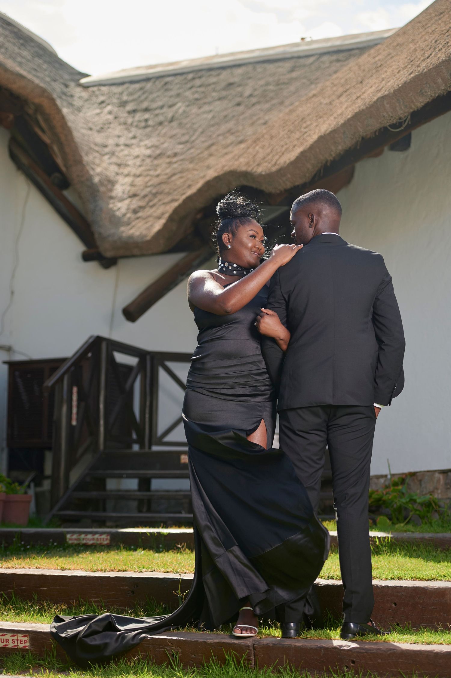 Elegant couple in matching black formal attire posing outside a thatched roof building on a sunny day.