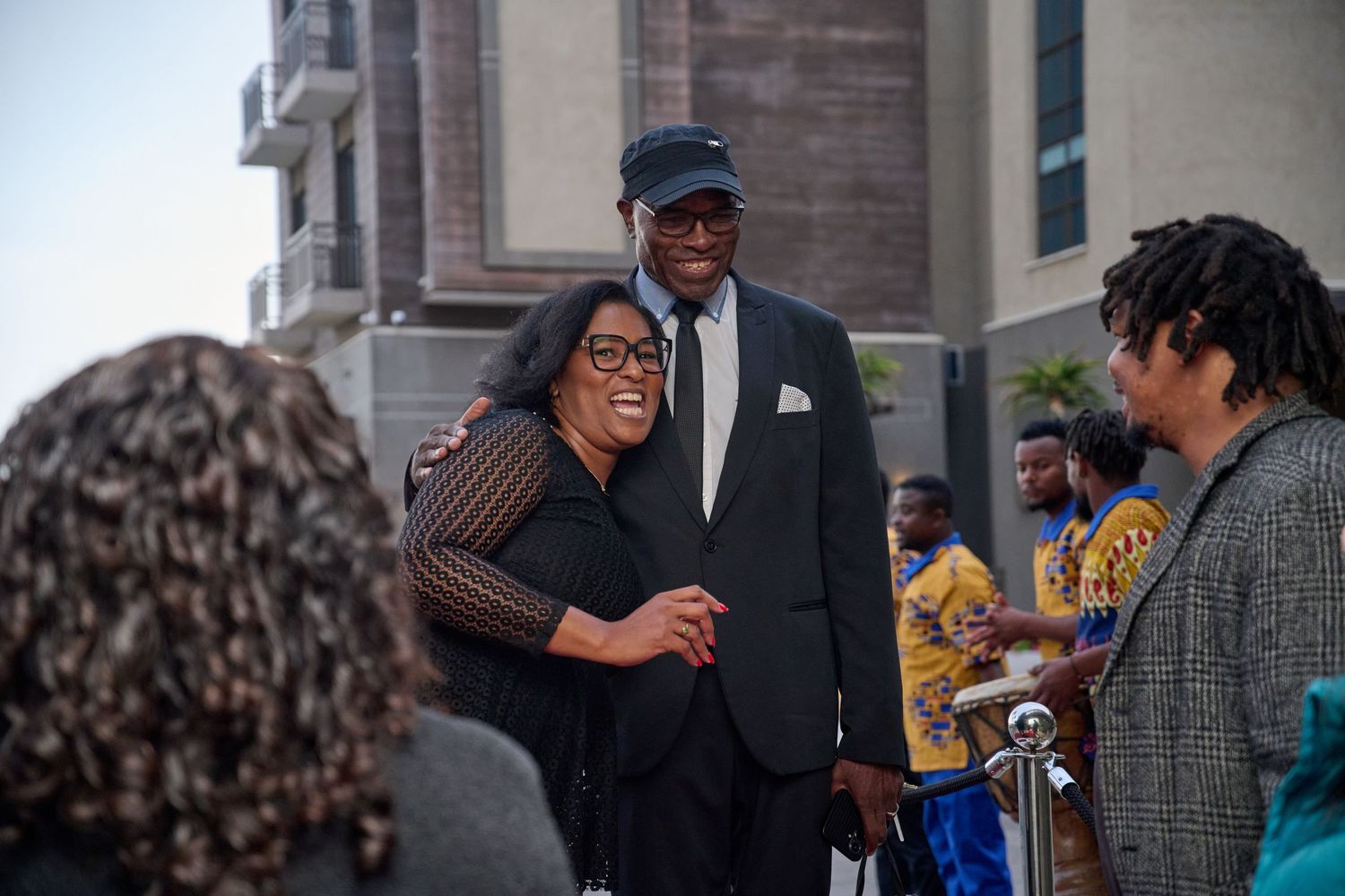 Tall man in suit smiling with woman at outdoor event surrounded by guests.