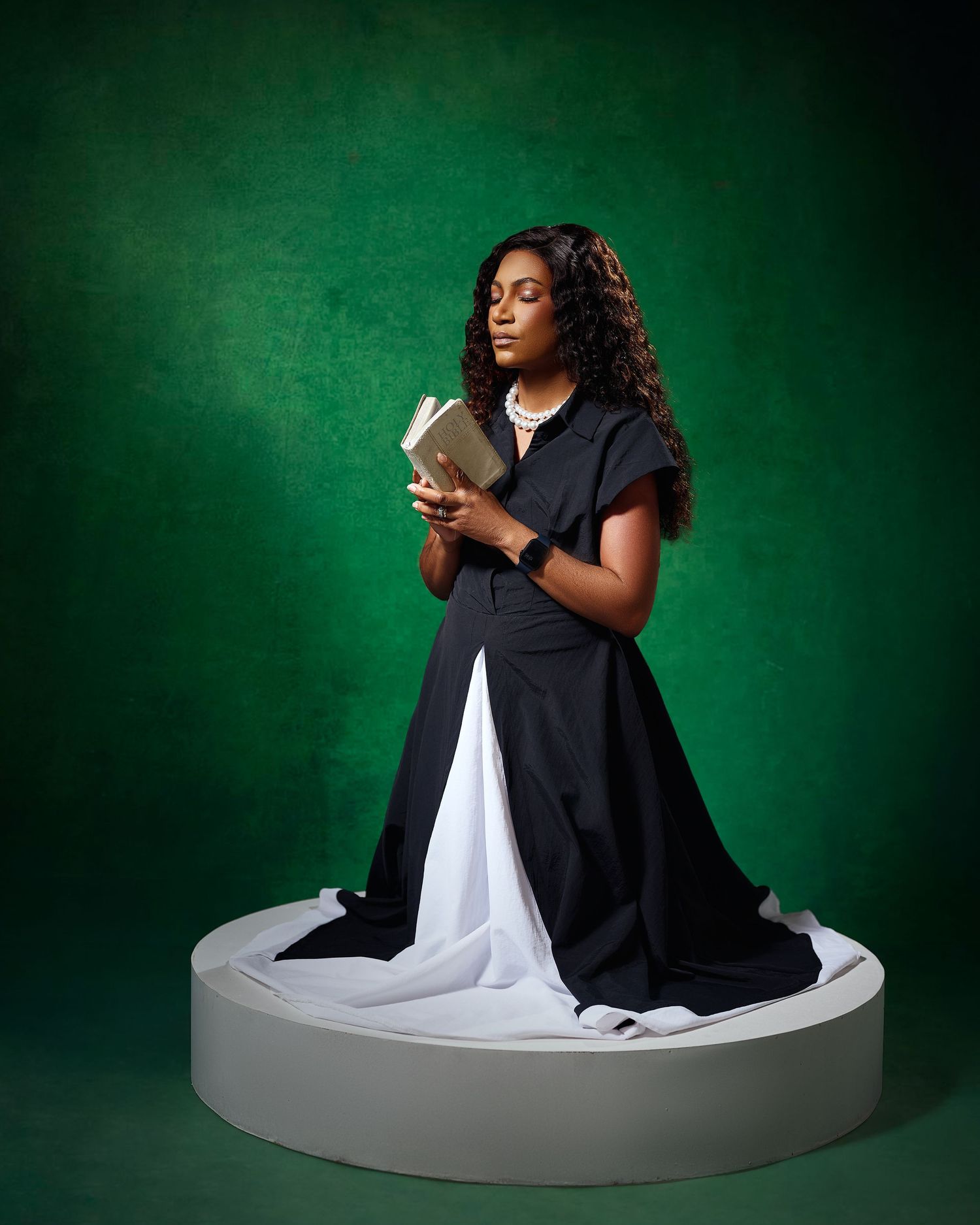 Woman in black dress reading a book, posed on white platform against green background.