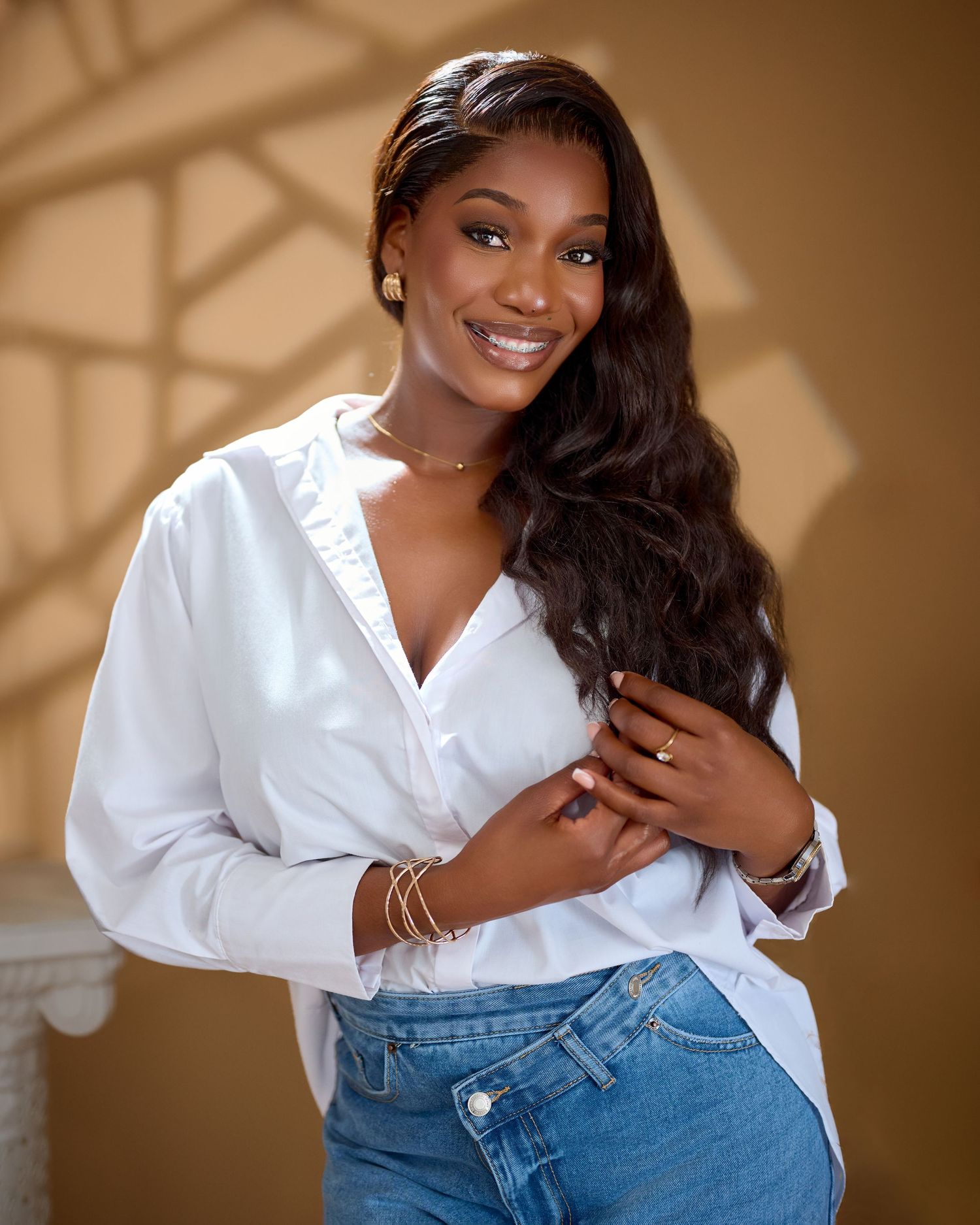 Smiling young woman in white blouse and jeans posing confidently with gold jewelry and long wavy hair.