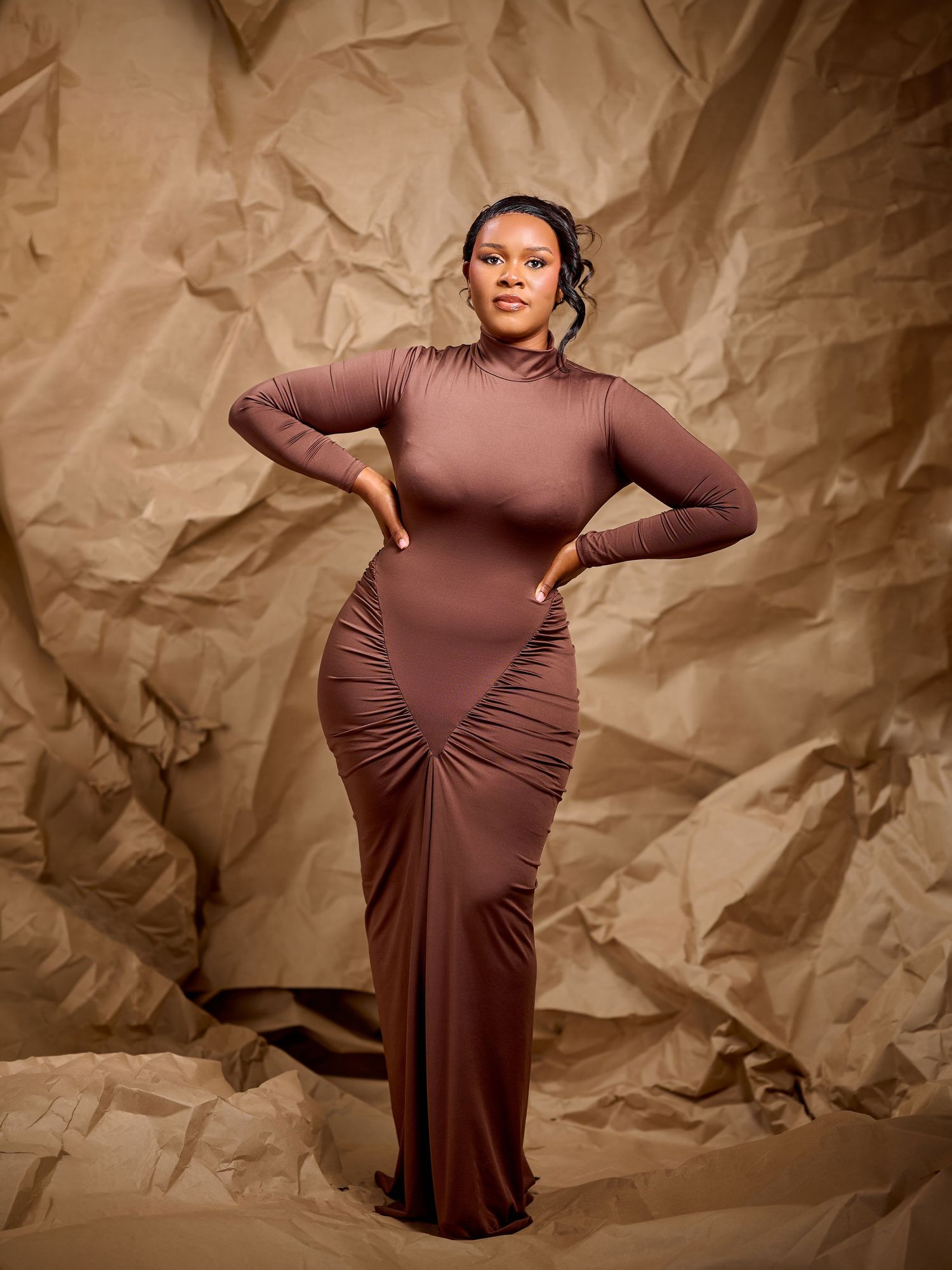 Woman in brown long-sleeve maxi dress posing confidently against crinkled brown paper background.