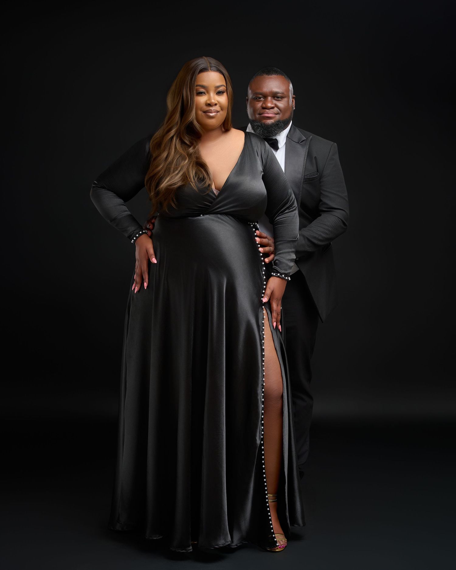 Elegant couple posing in matching black formal attire against a dark studio background.
