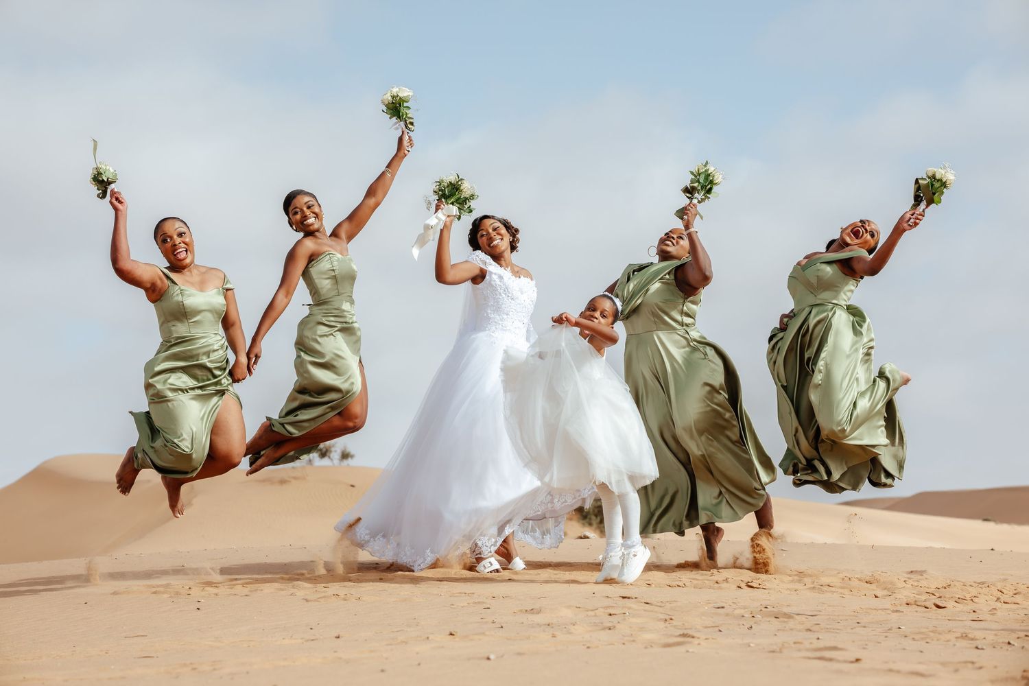 Bride and bridesmaids in sage green dresses joyfully jumping on golden sand dunes, holding bouquets.